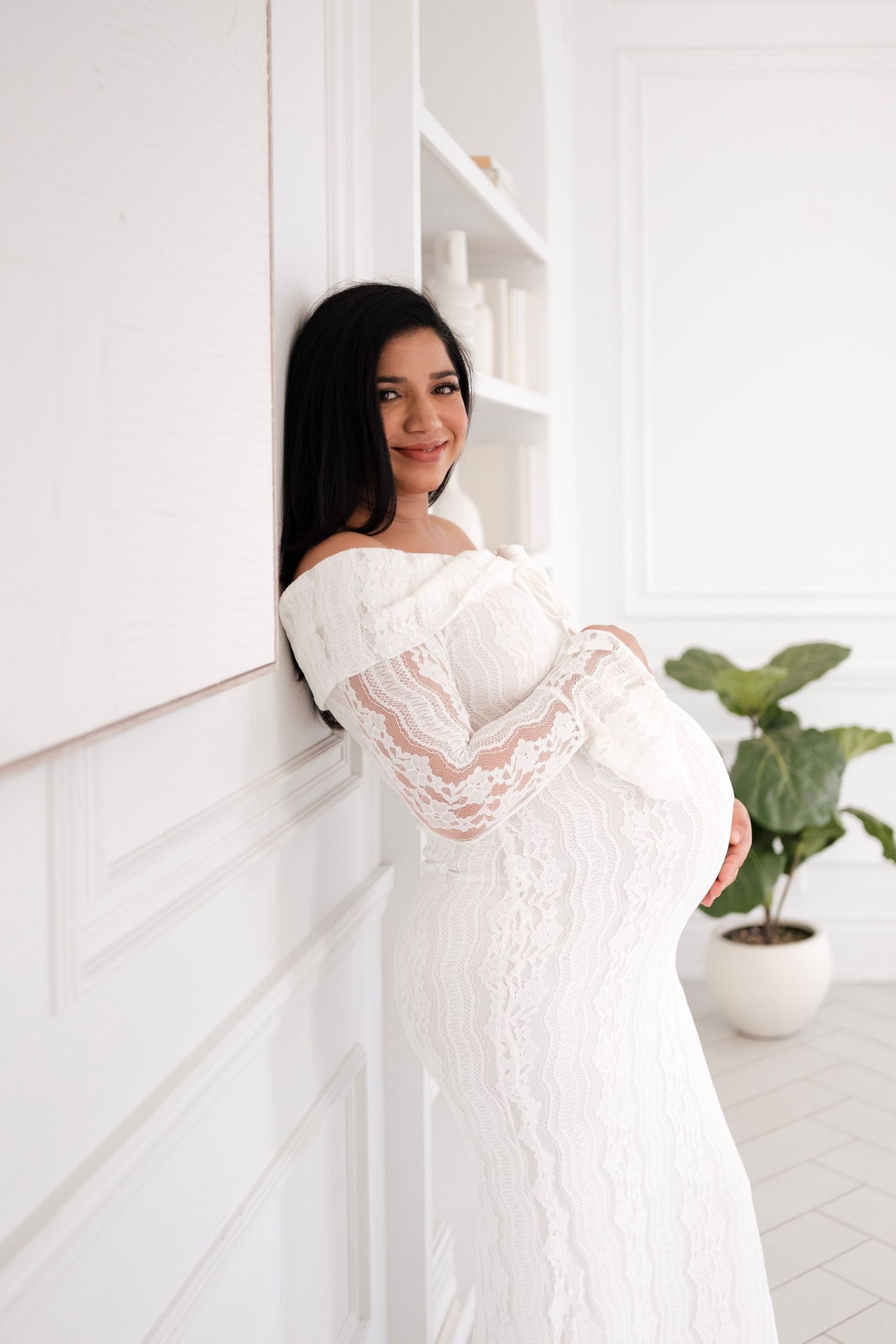 Smiling pregnant woman in white lace off-shoulder gown cradling baby bump, leaning against white wall.