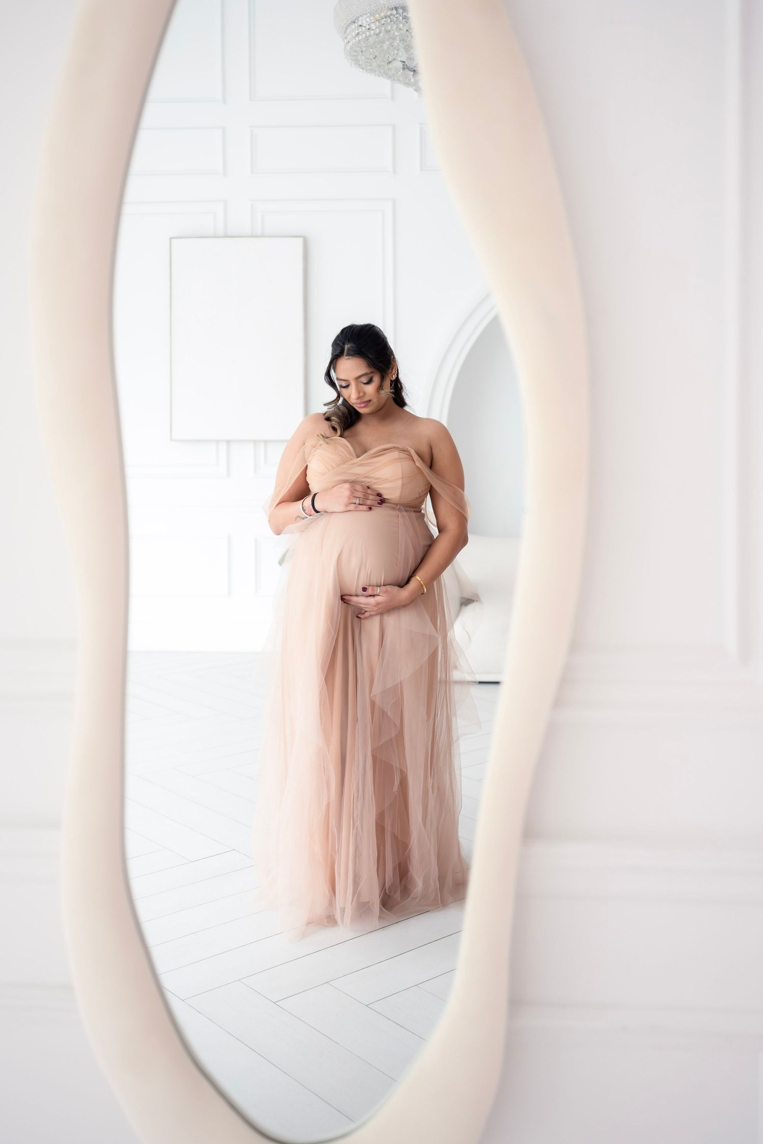 A pregnant woman in a blush pink tulle dress poses gracefully in a bright white studio setting.