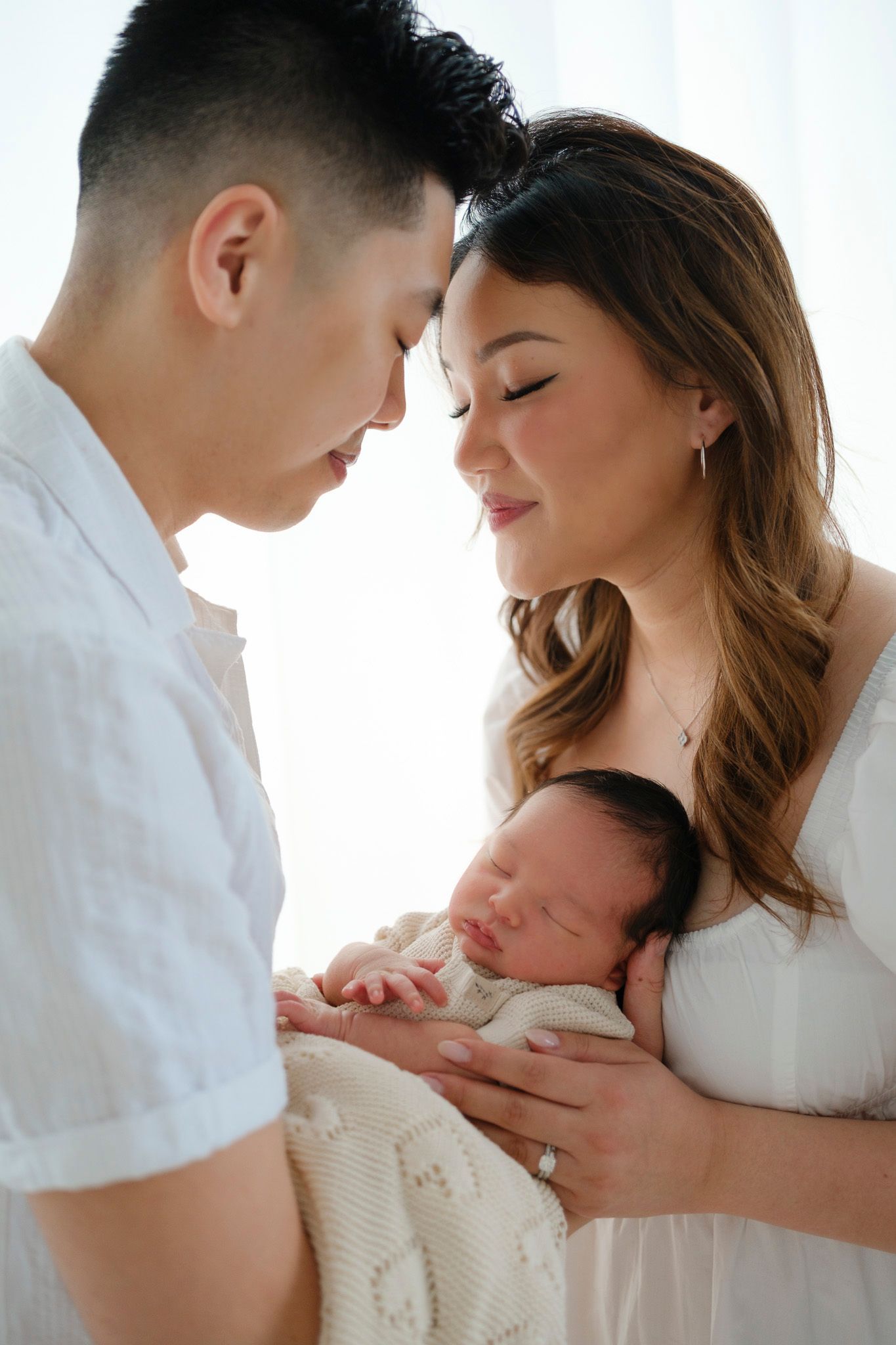 A tender family moment showing parents gazing lovingly at their sleeping newborn baby wrapped in cream colored blanket.