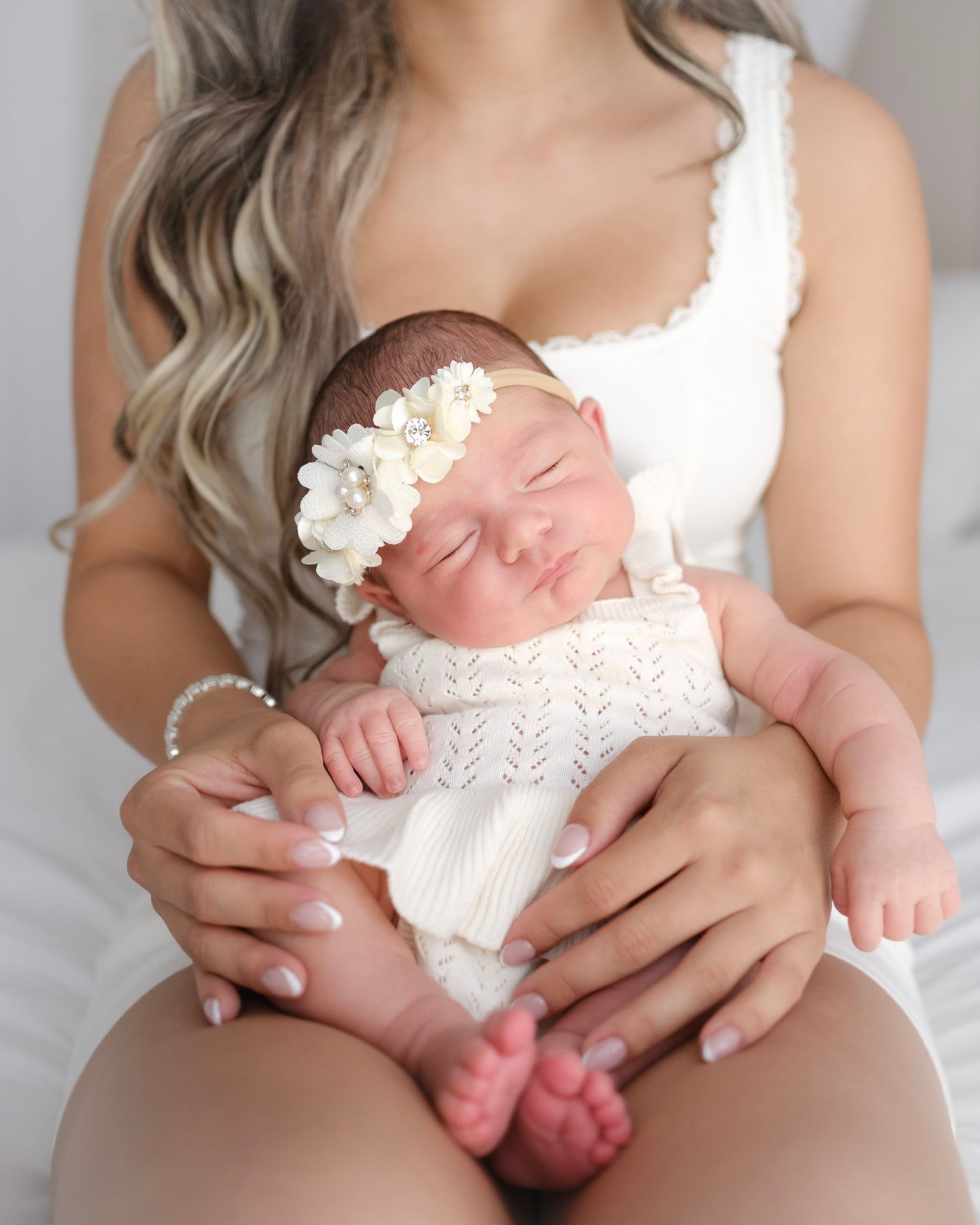 Mother in white dress cradling sleeping newborn baby wearing floral headband and knit white outfit.