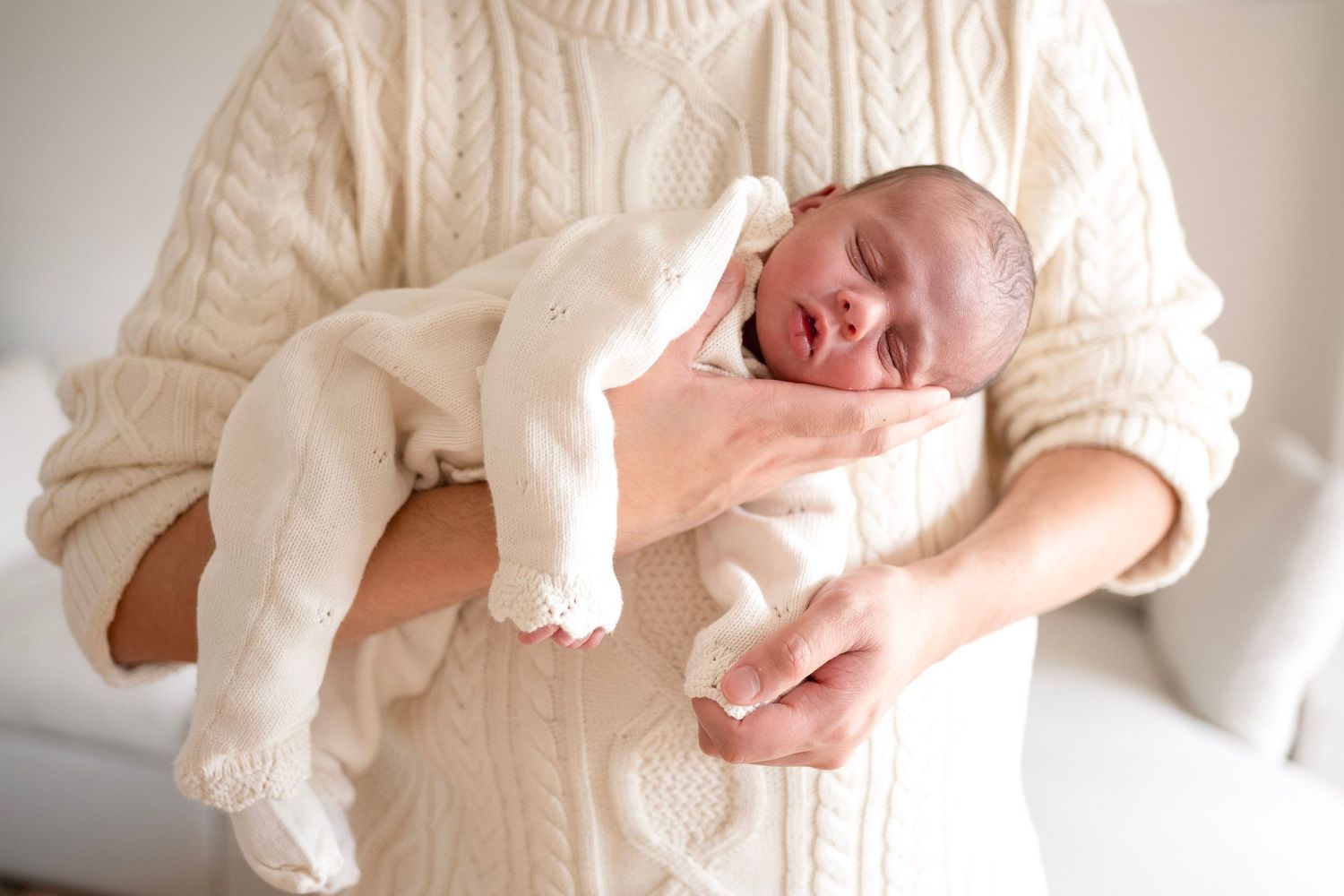 A newborn baby sleeping peacefully in cream outfit, cradled in the arms of a person wearing a white cable-knit sweater.