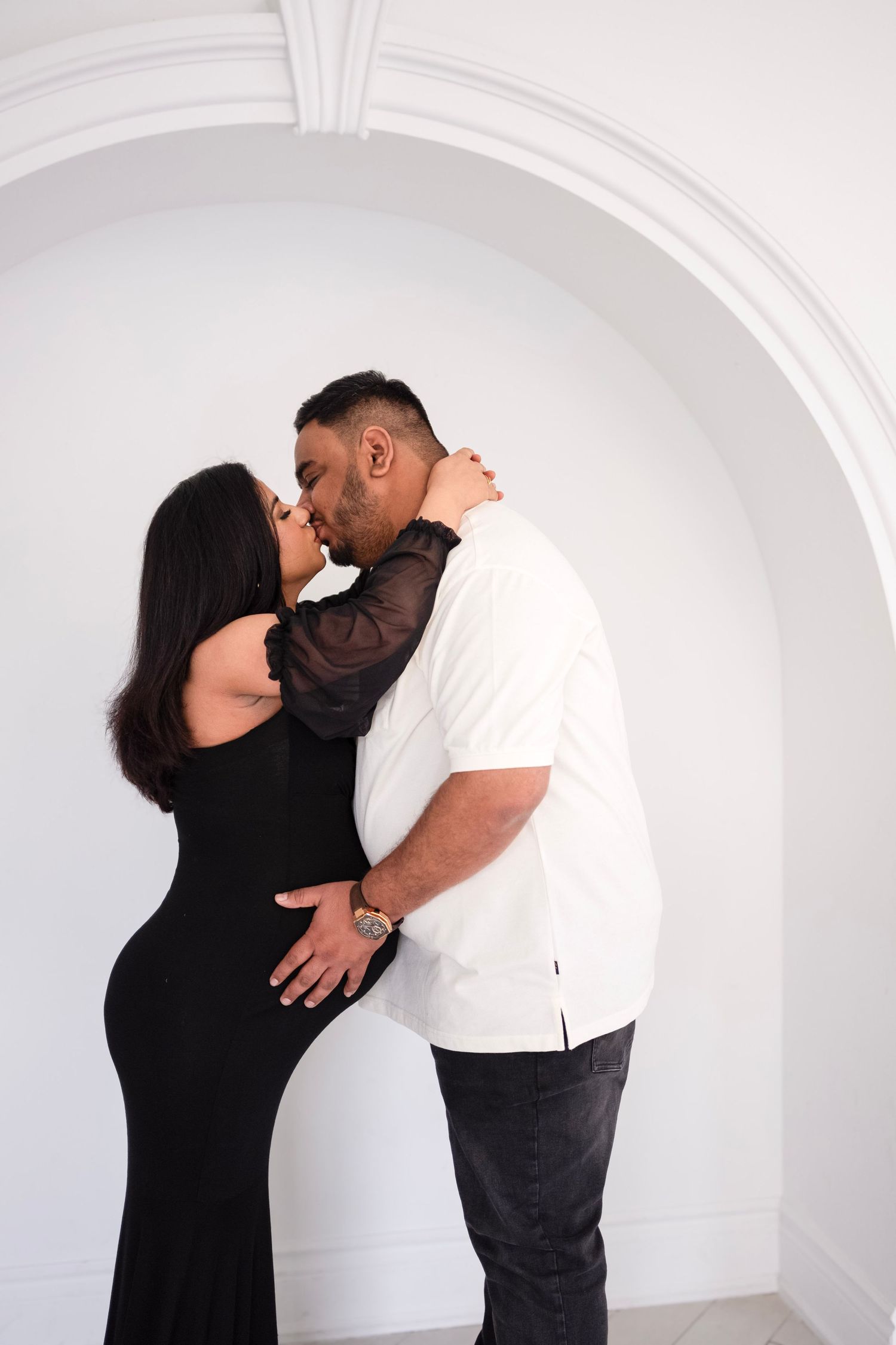 Couple sharing a romantic kiss under a white arched doorway, woman in black dress, man in white shirt.