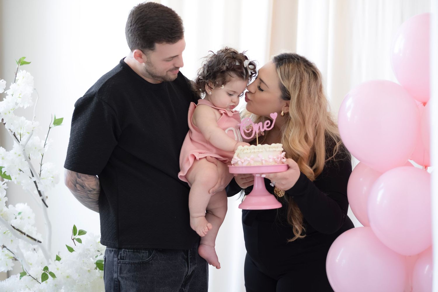 A family celebrates their baby girl's first birthday with a pink cake and balloon decorations.