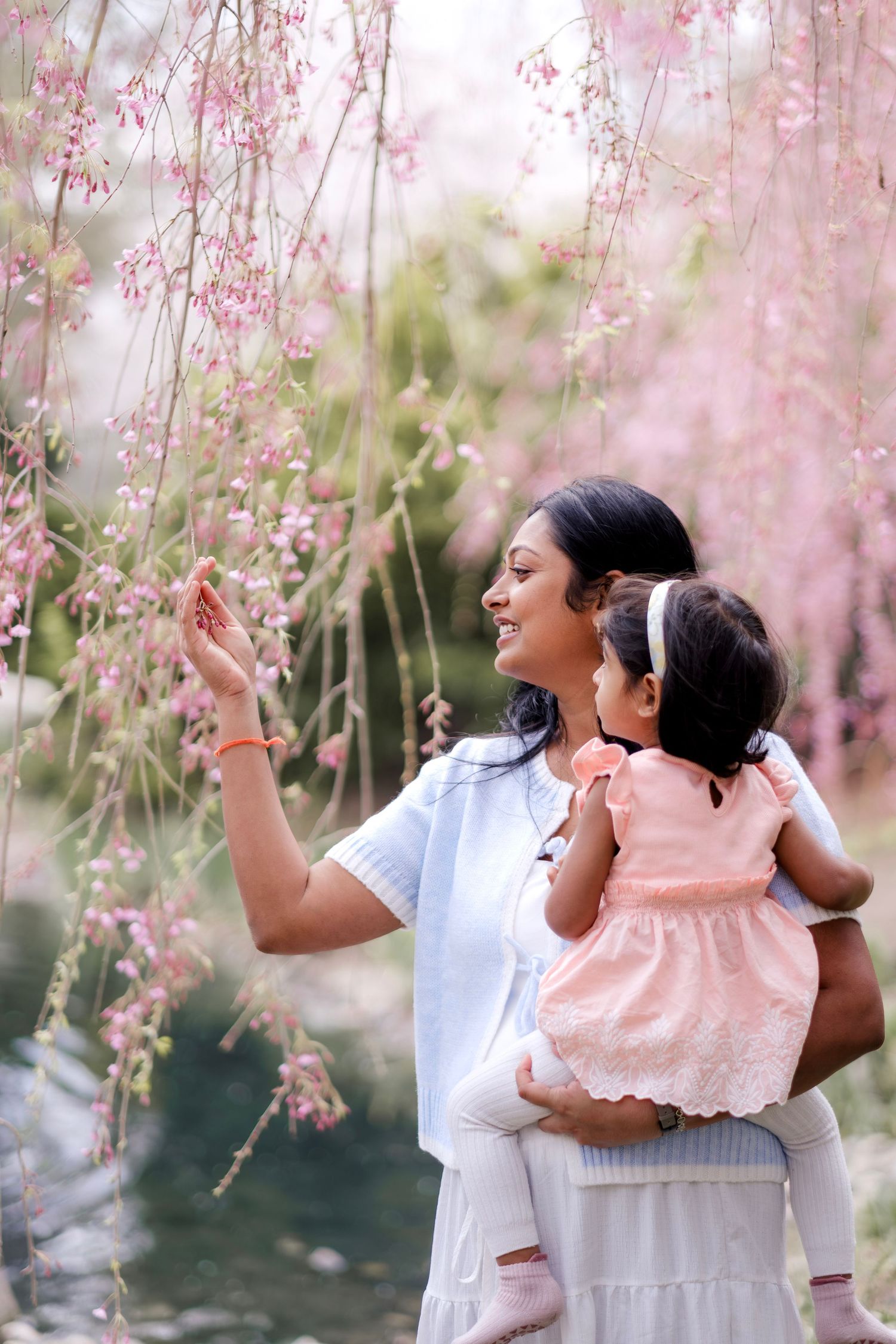 A mother holds her baby girl among pink cherry blossom branches, both dressed in light colors, smiling outdoors.