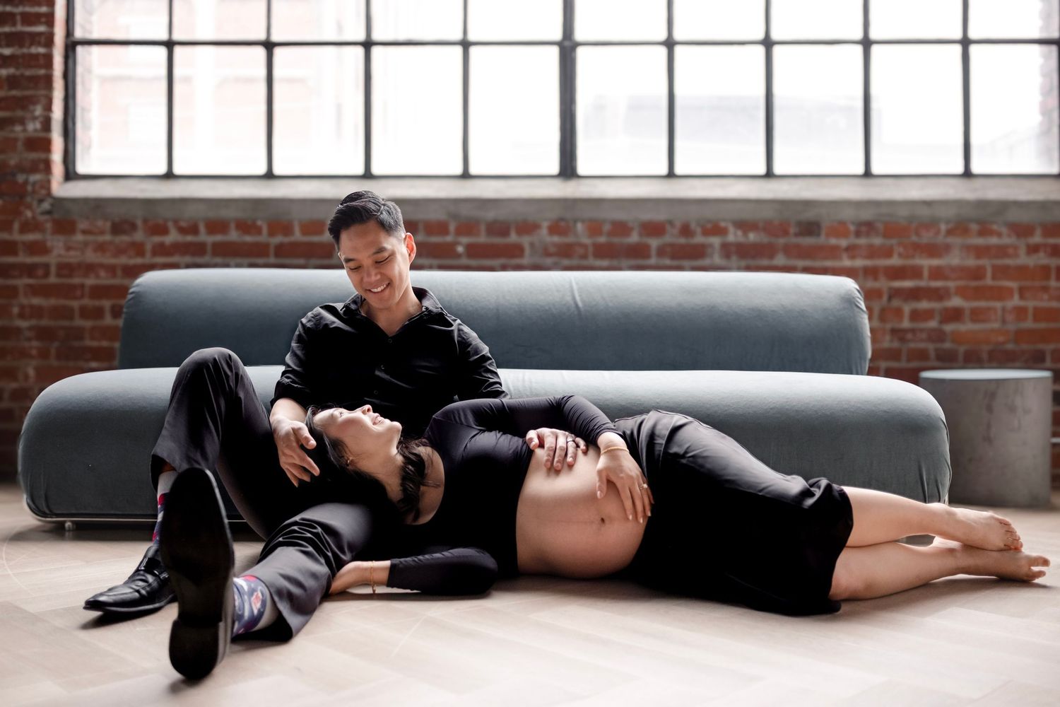 Couple posing on floor in black outfits, pregnant woman lying down, brick wall loft background.