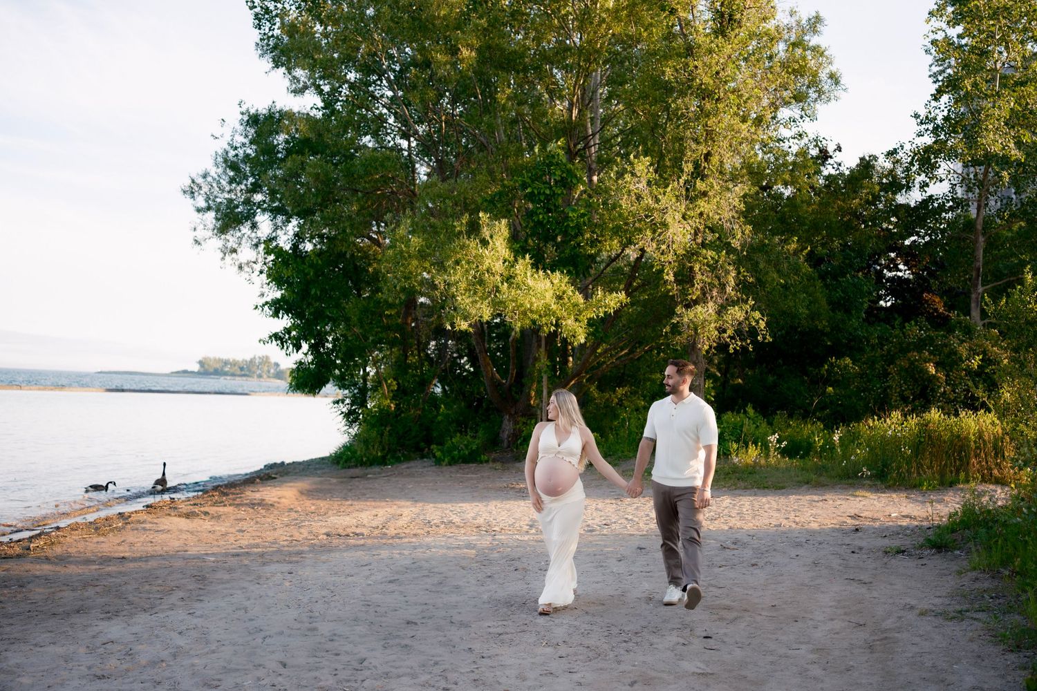Couple in wedding attire holding hands walking along a sandy beach shoreline surrounded by lush green trees.
