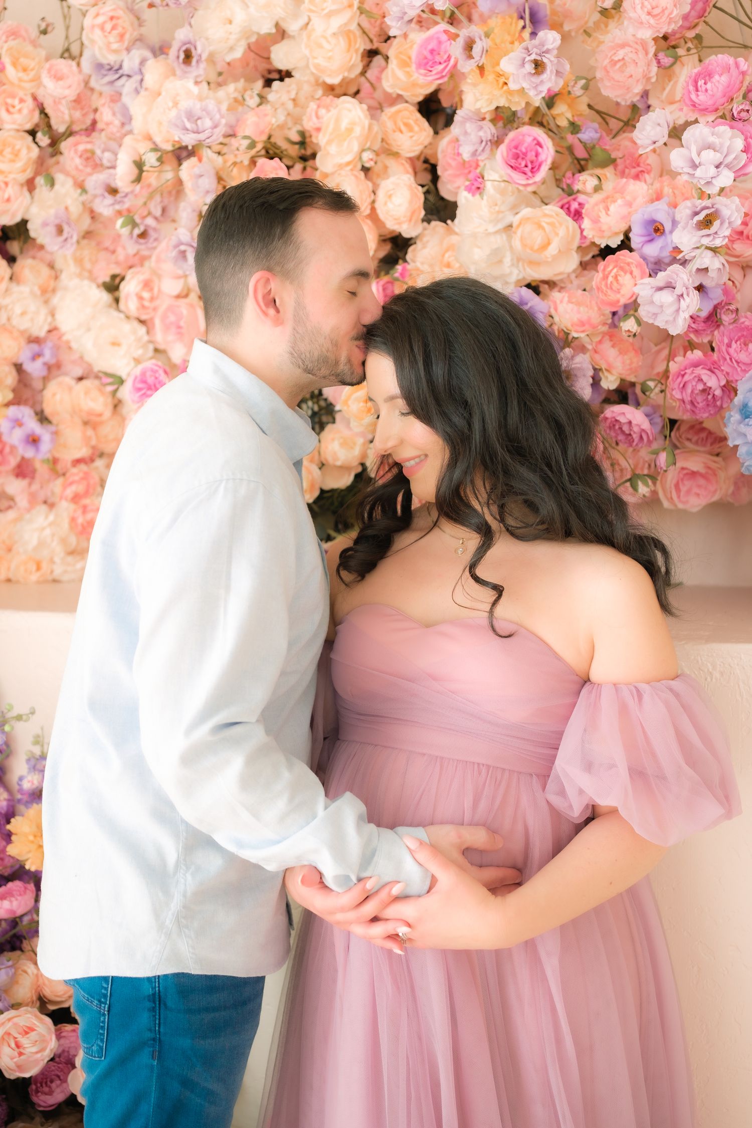 A couple shares an intimate moment in front of a romantic floral wall backdrop of pink and peach roses.