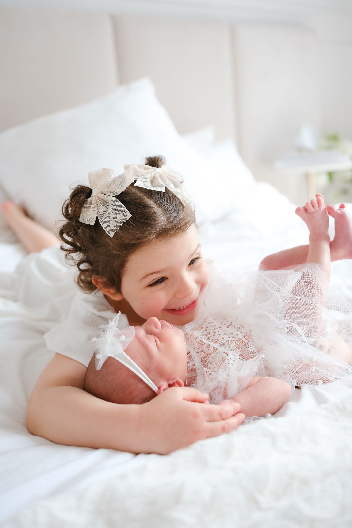 A joyful young child wearing a white lacy dress and bows lies on plush white bedding while smiling and playing.