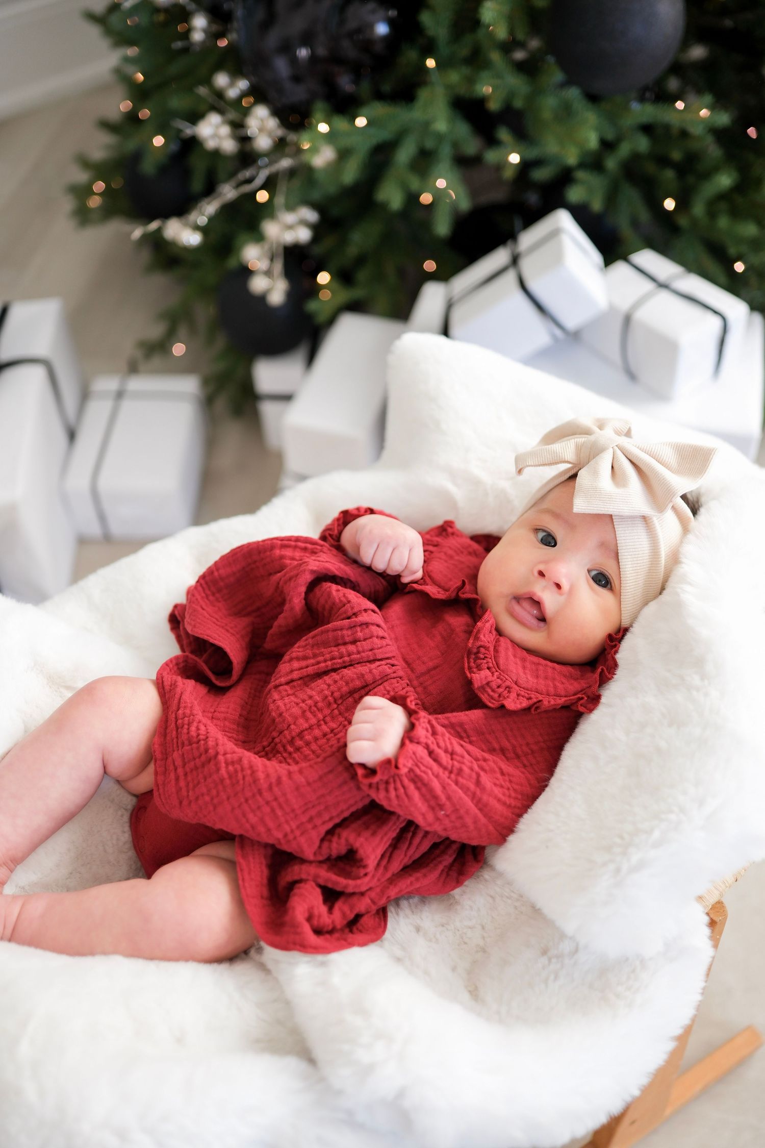 A baby wearing a red knit sweater lies on a white blanket with Christmas decorations in the background.