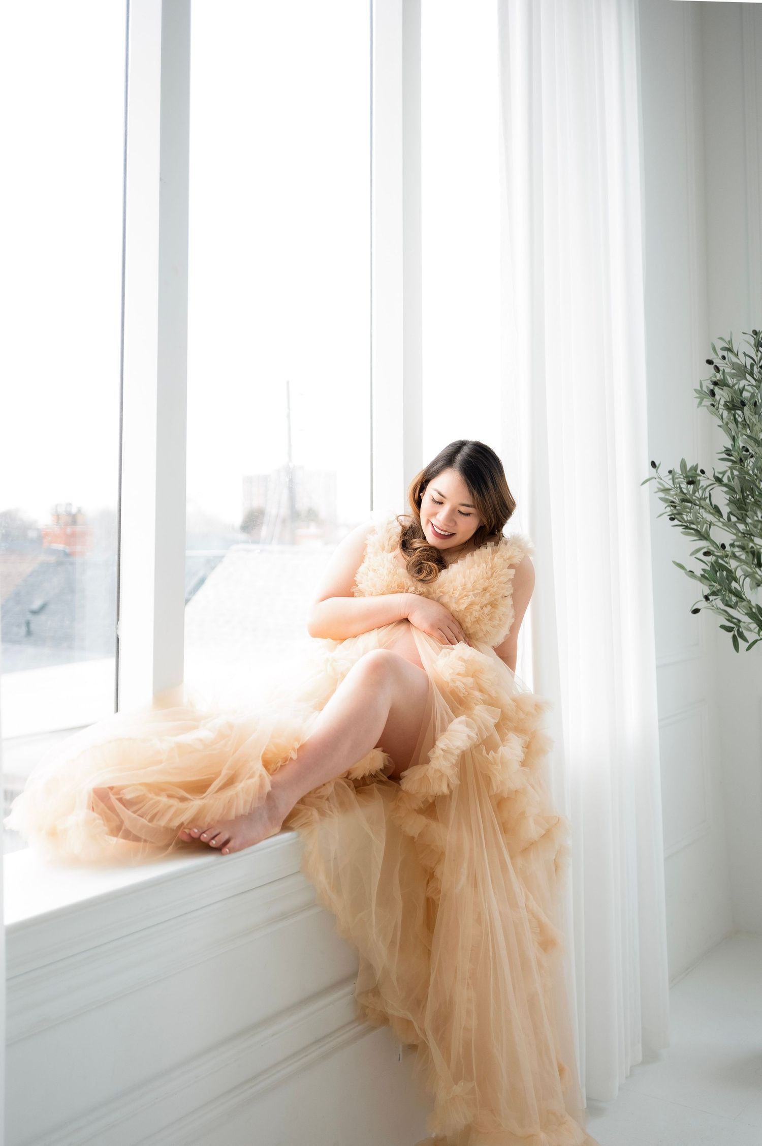 A person in a flowing peach tulle gown reclines gracefully against white windowsills in a bright, airy space.