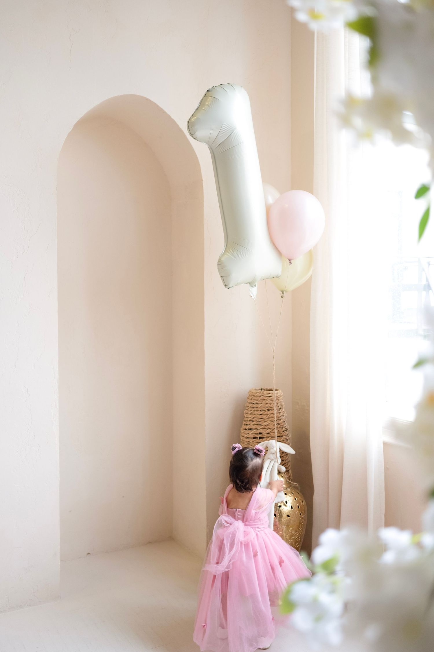 A toddler girl in a pink tutu dress reaches for a number one balloon at her first birthday party celebration.
