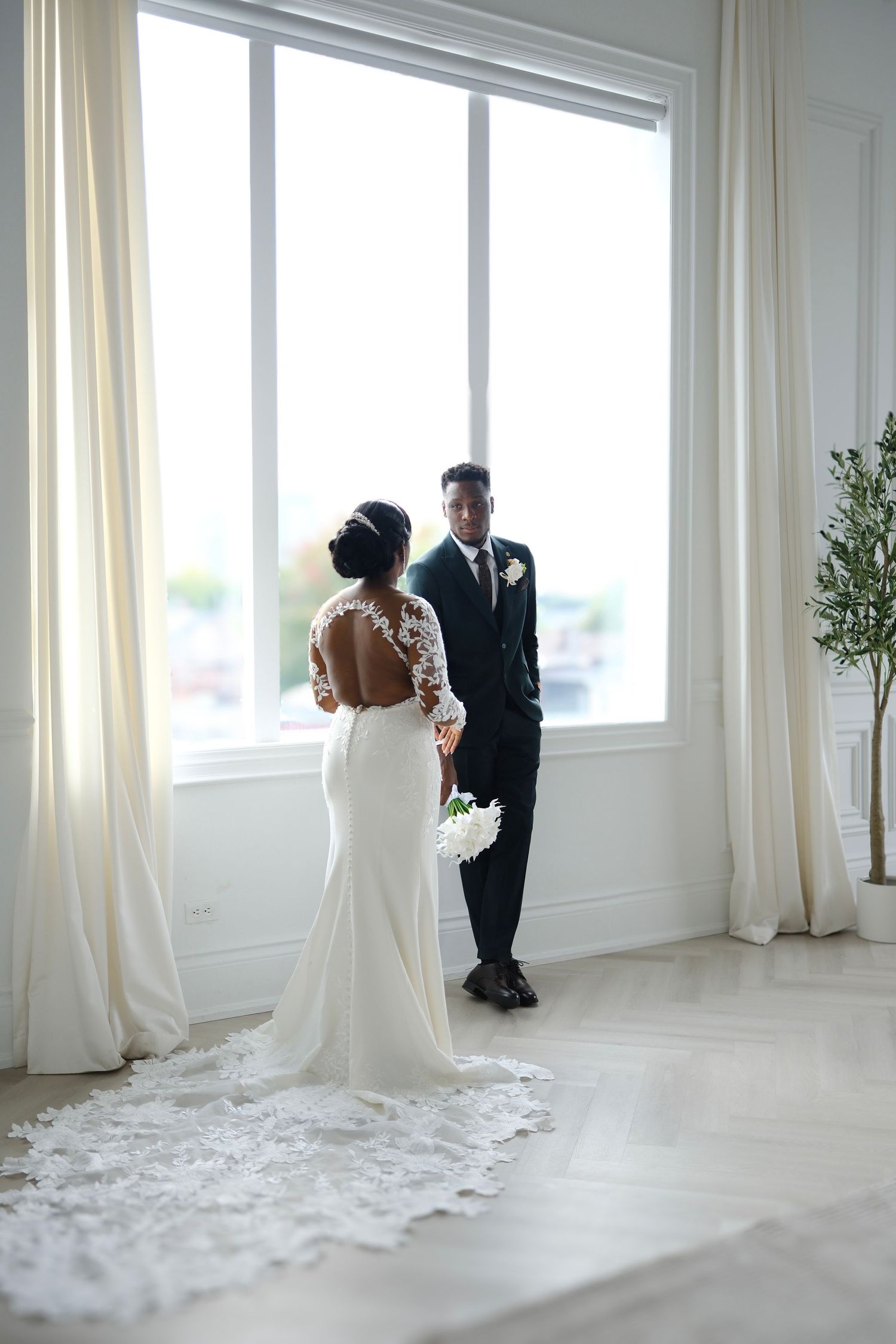 A romantic wedding scene with an elegant backless lace gown and black suit silhouetted against a bright window.