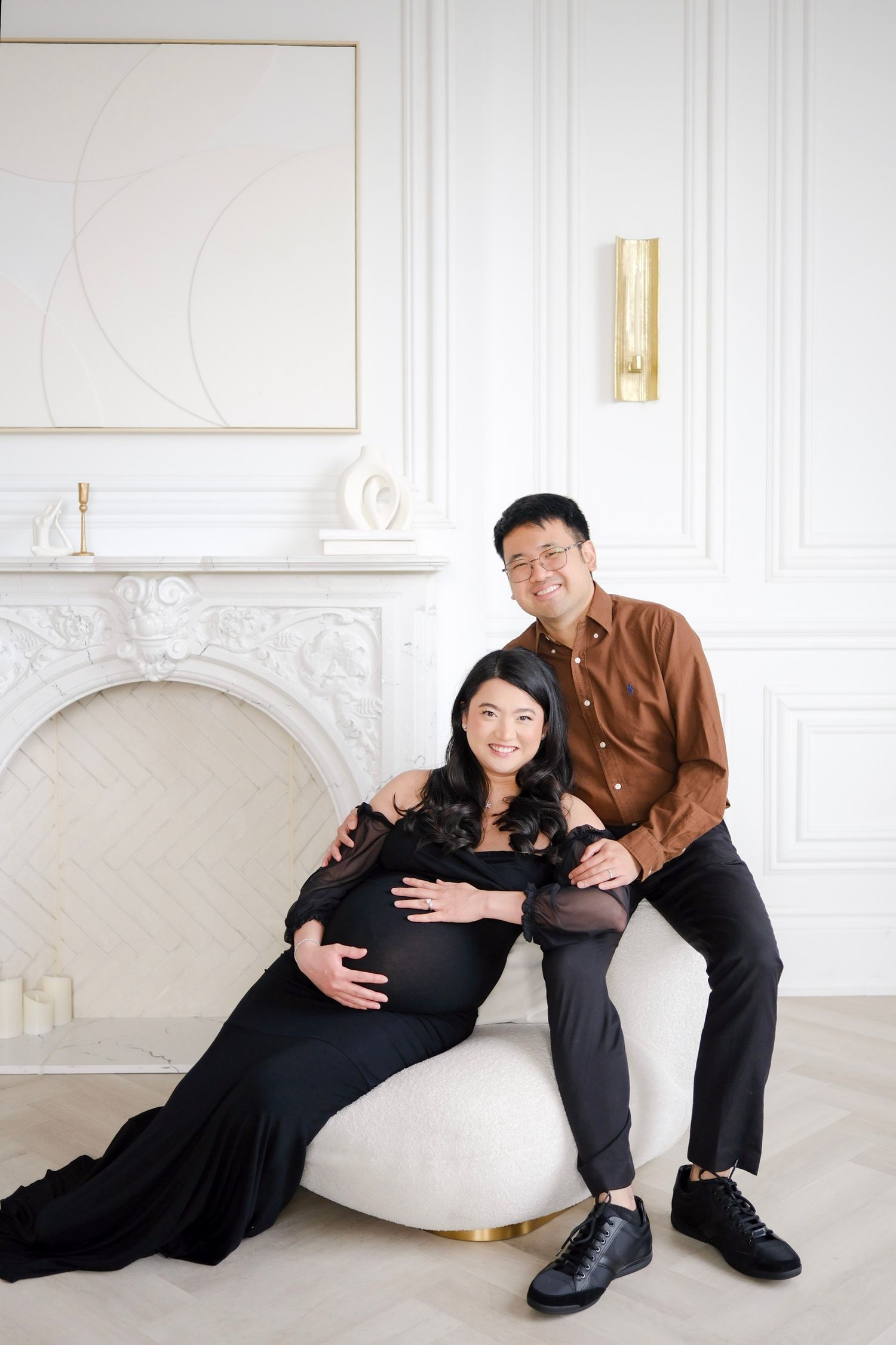 A couple poses together in an elegant white room with a marble fireplace, featuring a maternity photo session.