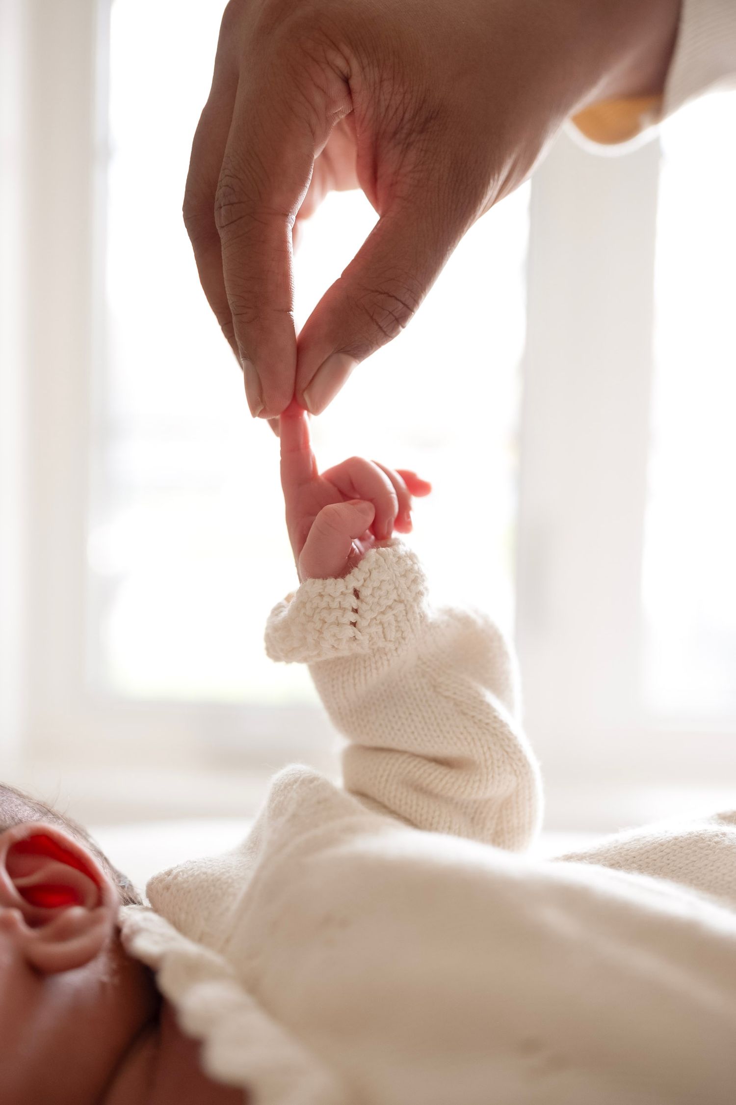 A parent's finger gently held by a newborn baby's tiny hand wearing a white knit mitten.
