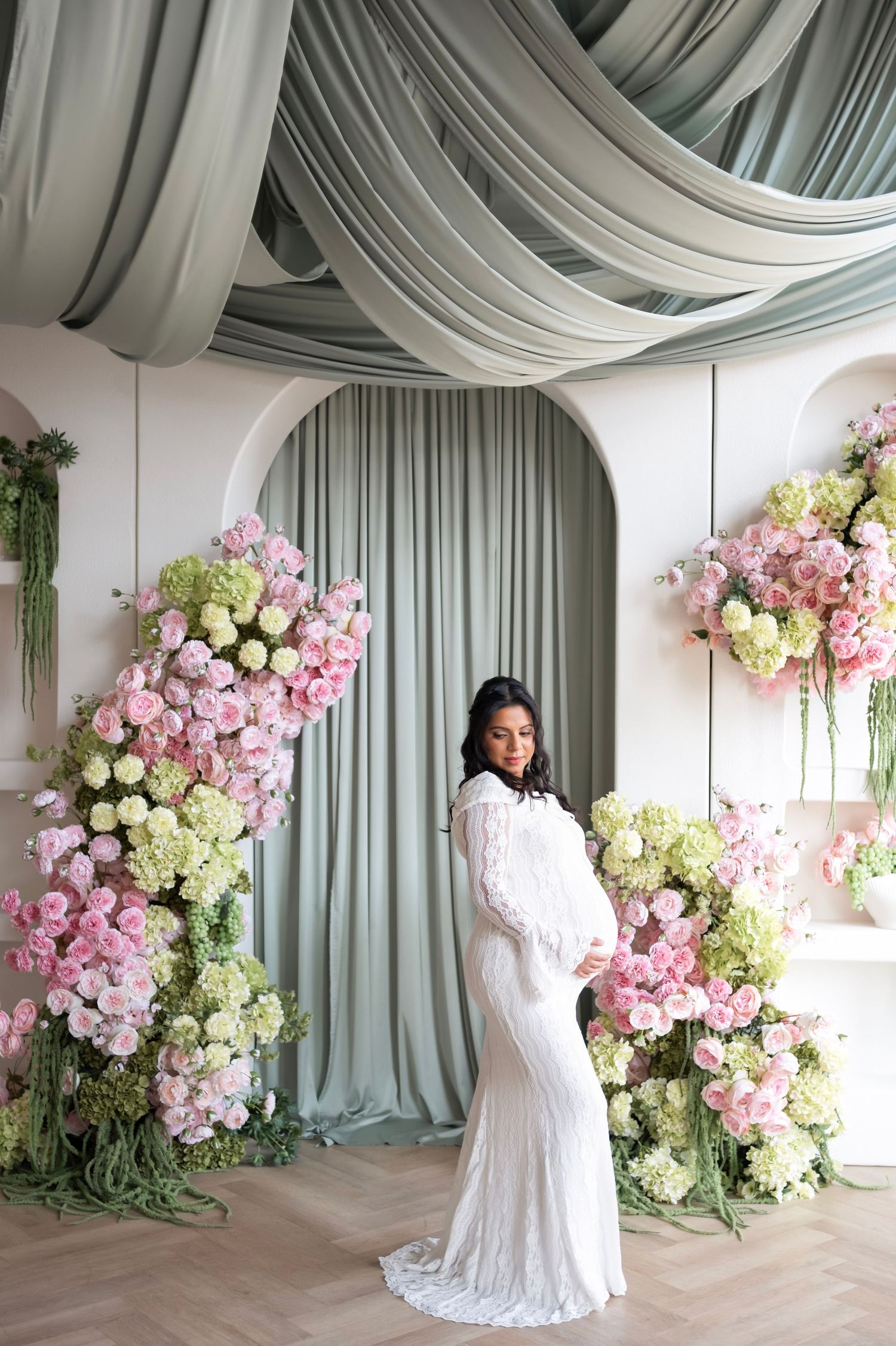 Pregnant woman in white lace gown poses before floral arch with pink roses and green hydrangeas.