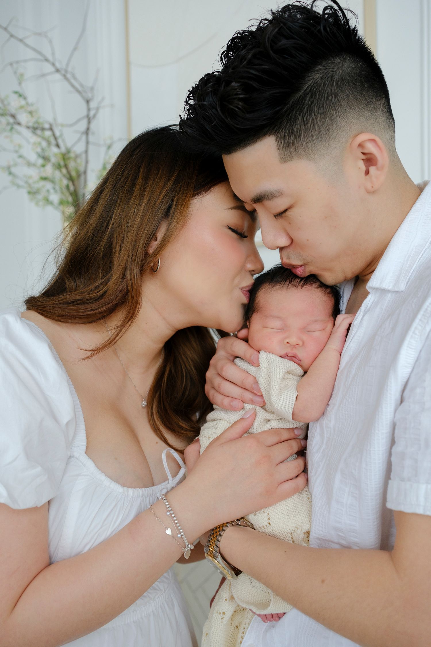A tender moment as parents in white outfits lovingly embrace their sleeping newborn baby between them.