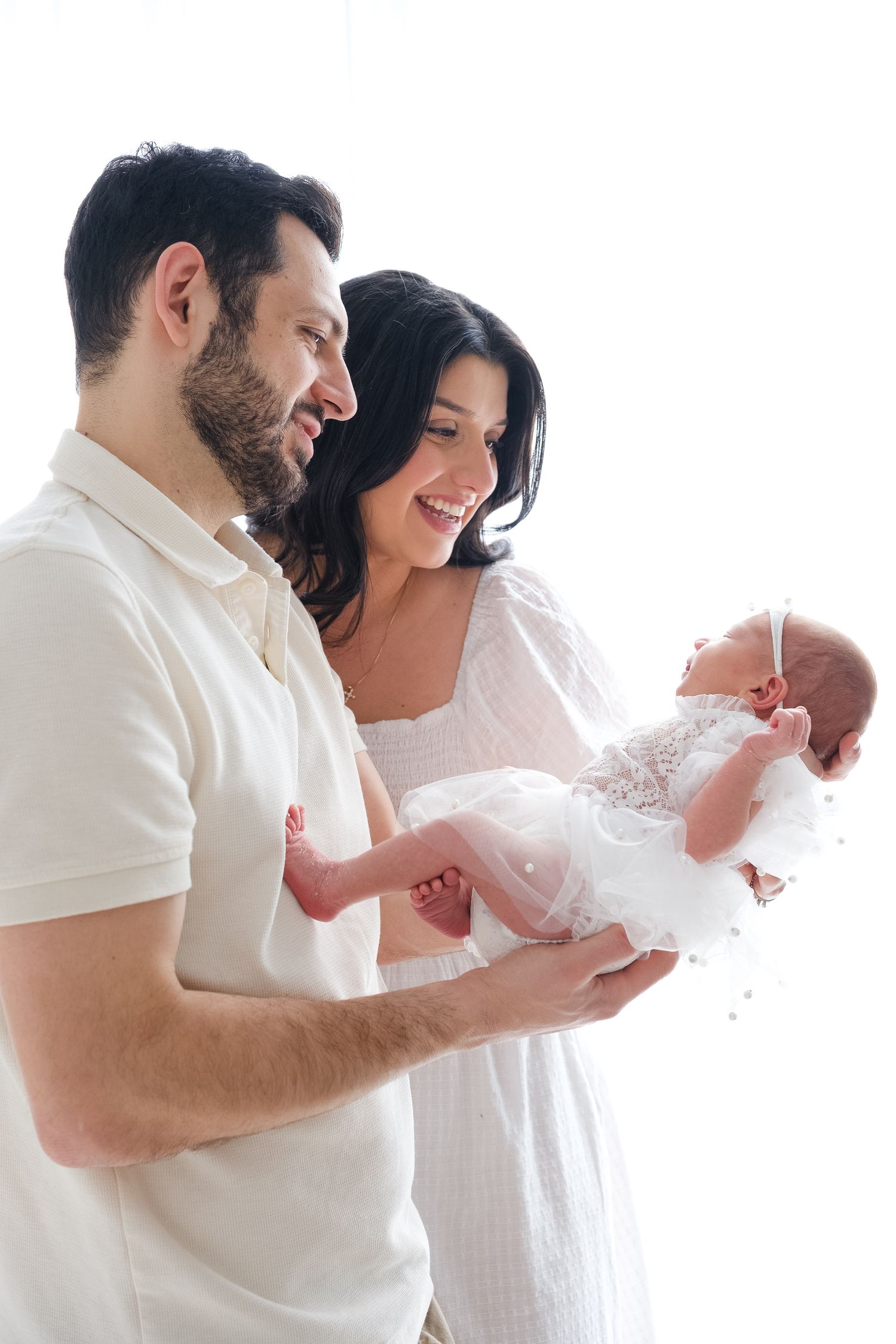 A couple in white clothing hold their newborn baby dressed in delicate lace against a bright background.