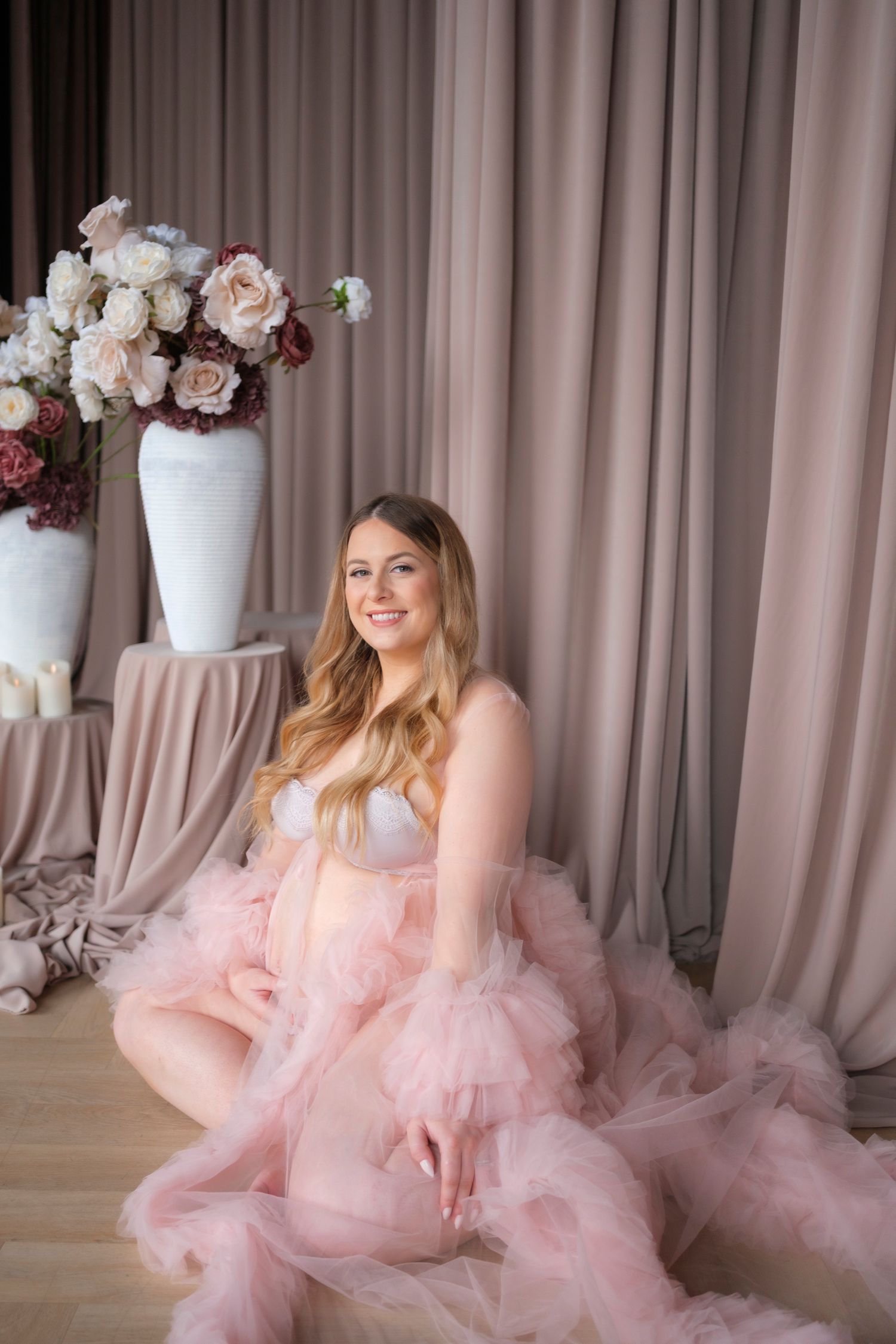 A dreamy portrait with someone wearing a flowing pink tulle gown sitting beside elegant floral arrangements in a studio setting.