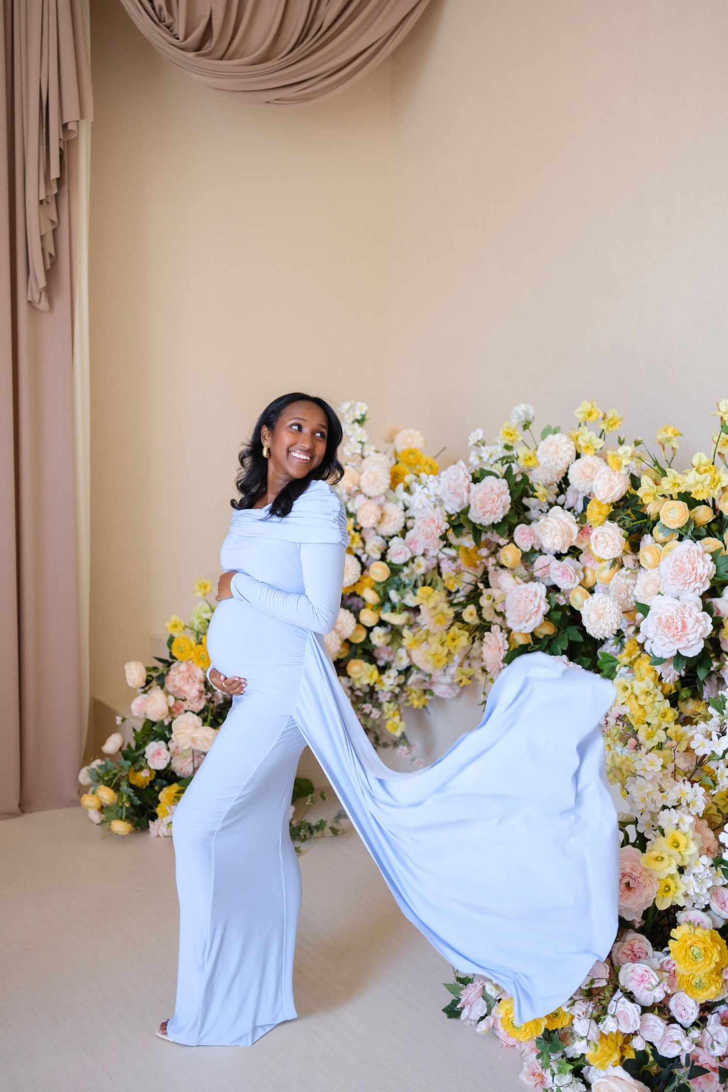 Pregnant woman in light blue gown posing in front of colorful floral arrangement during maternity photoshoot.