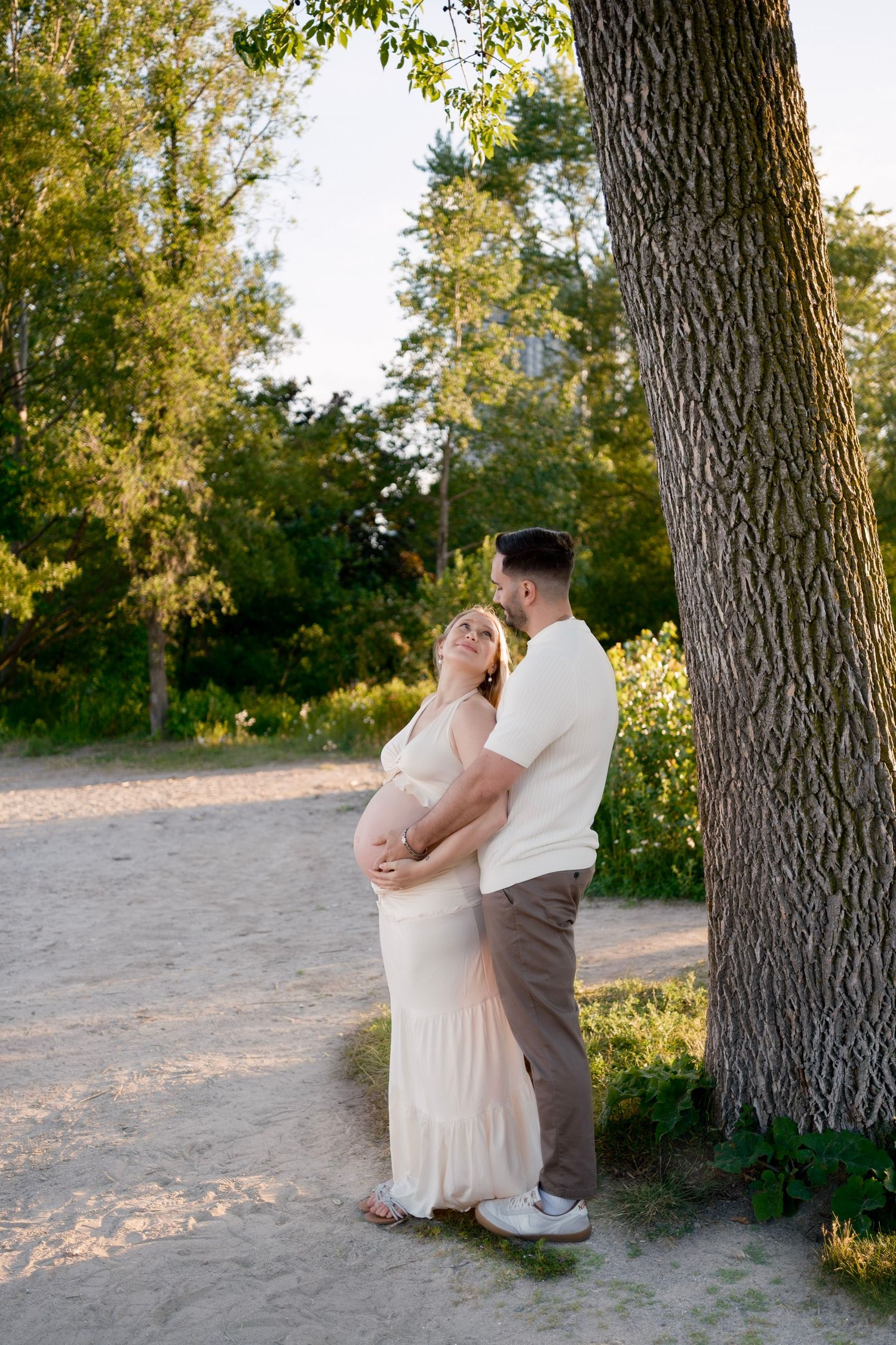 Pregnant couple embracing tenderly near a large tree on a sandy path in golden hour sunlight.