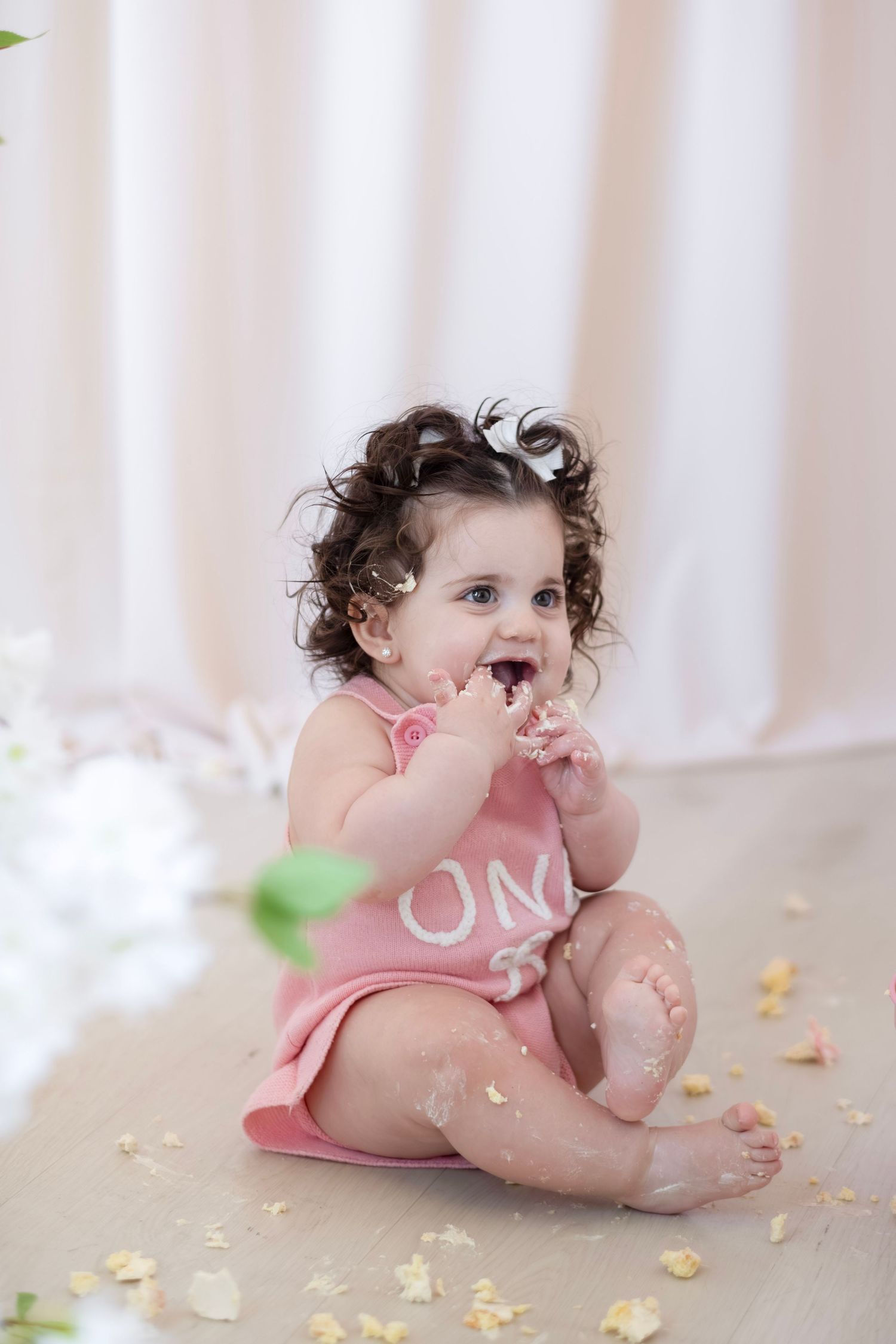 Curly-haired baby girl in pink romper sitting on floor smashing first birthday cake, with frosting on her face and hands.