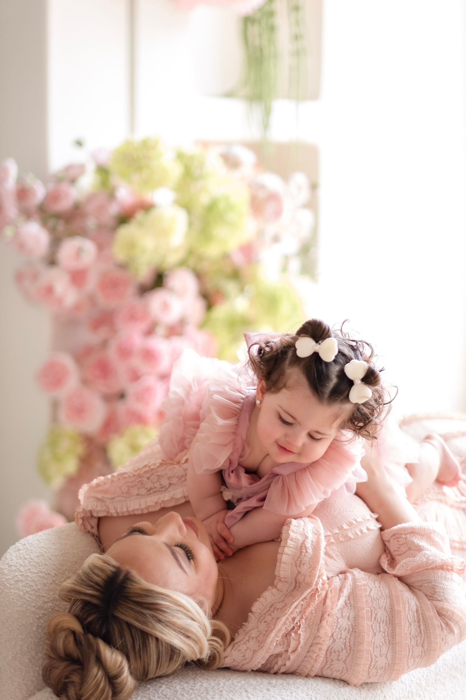 Mother lying down with young daughter in pink dresses surrounded by soft pink and green floral arrangement.