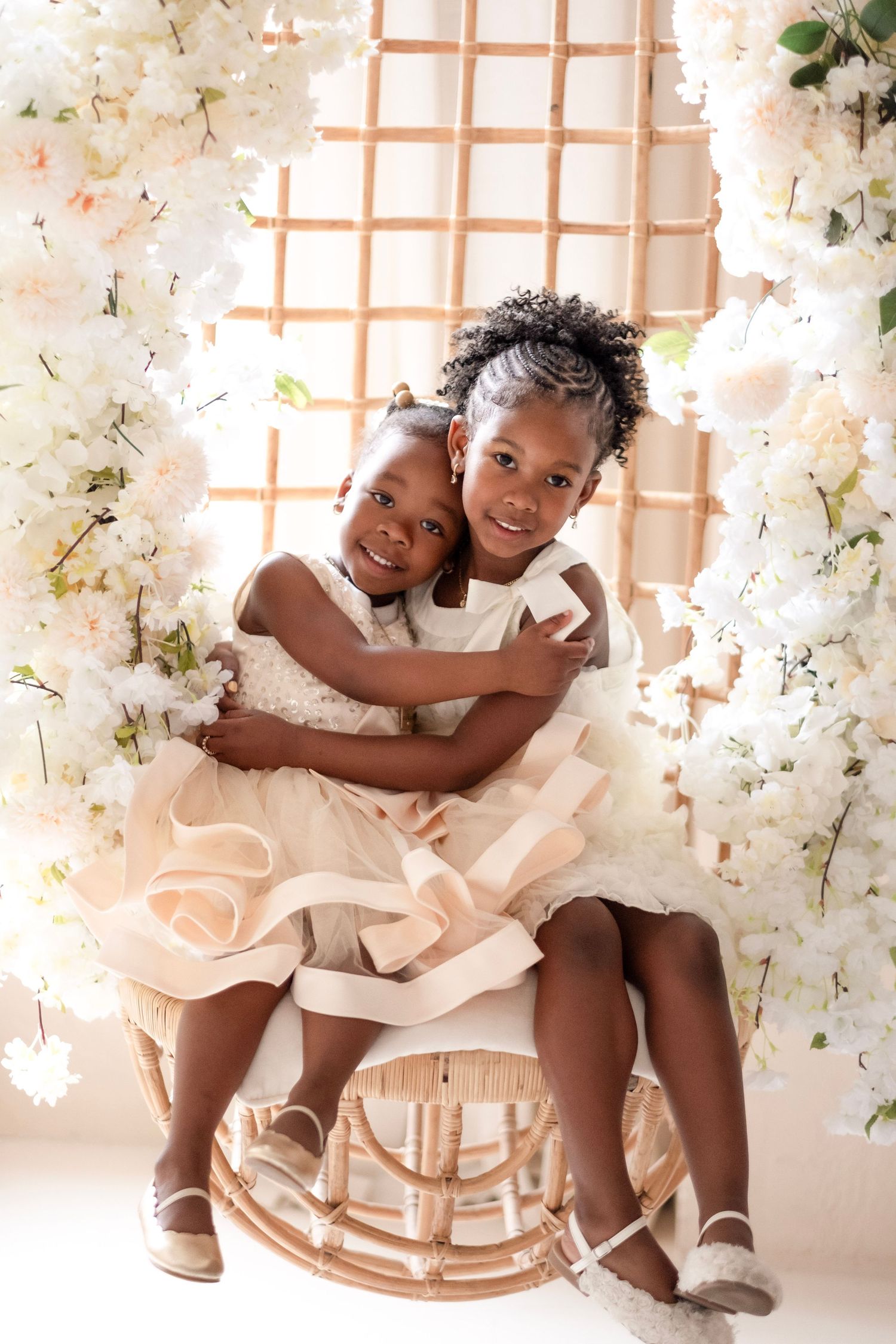 Two young Black girls in elegant dresses share a warm hug on a rattan chair with white florals.