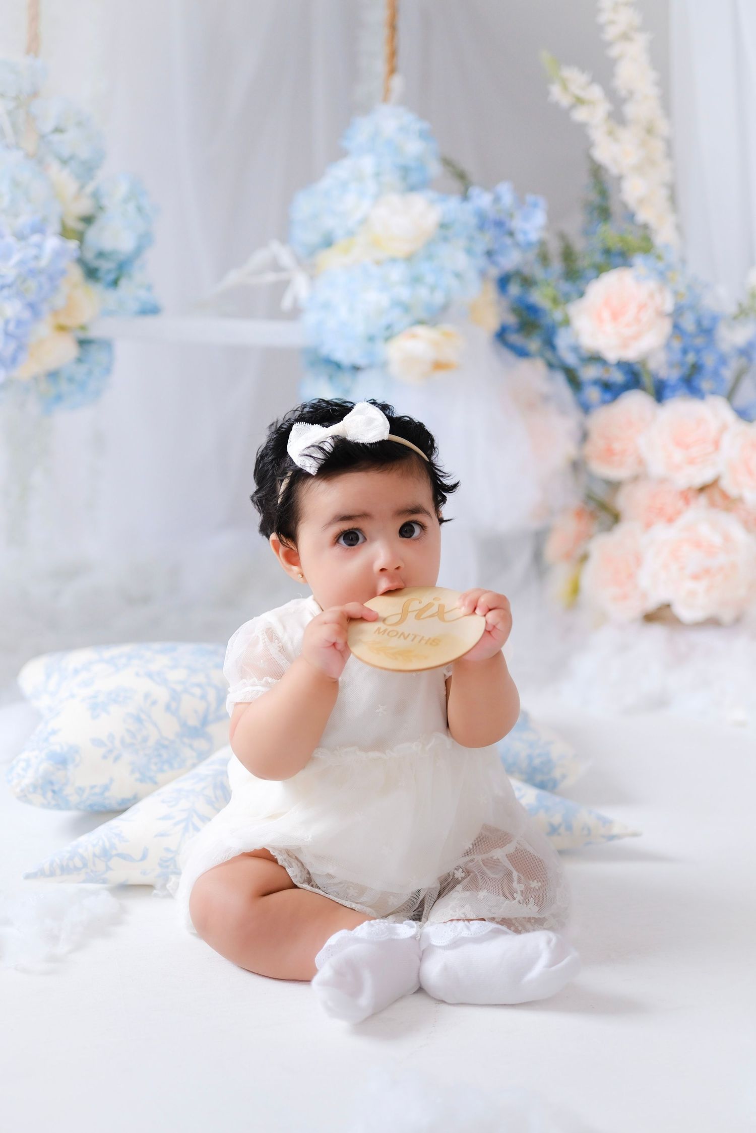 Sweet baby in white dress sits among pastel blue and pink floral decorations while holding a cookie.