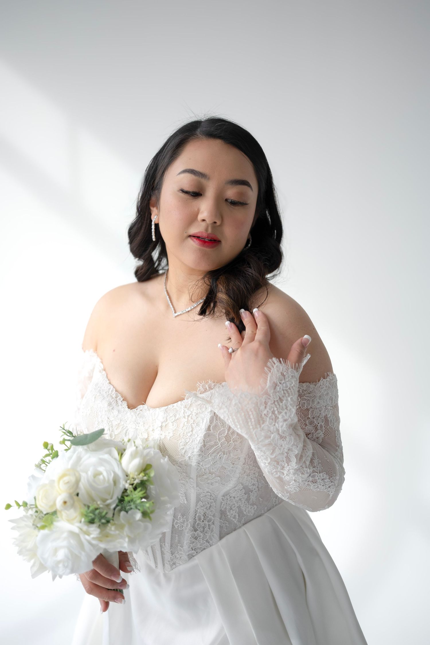 Asian bride in off-shoulder lace wedding gown holds white floral bouquet against white background.