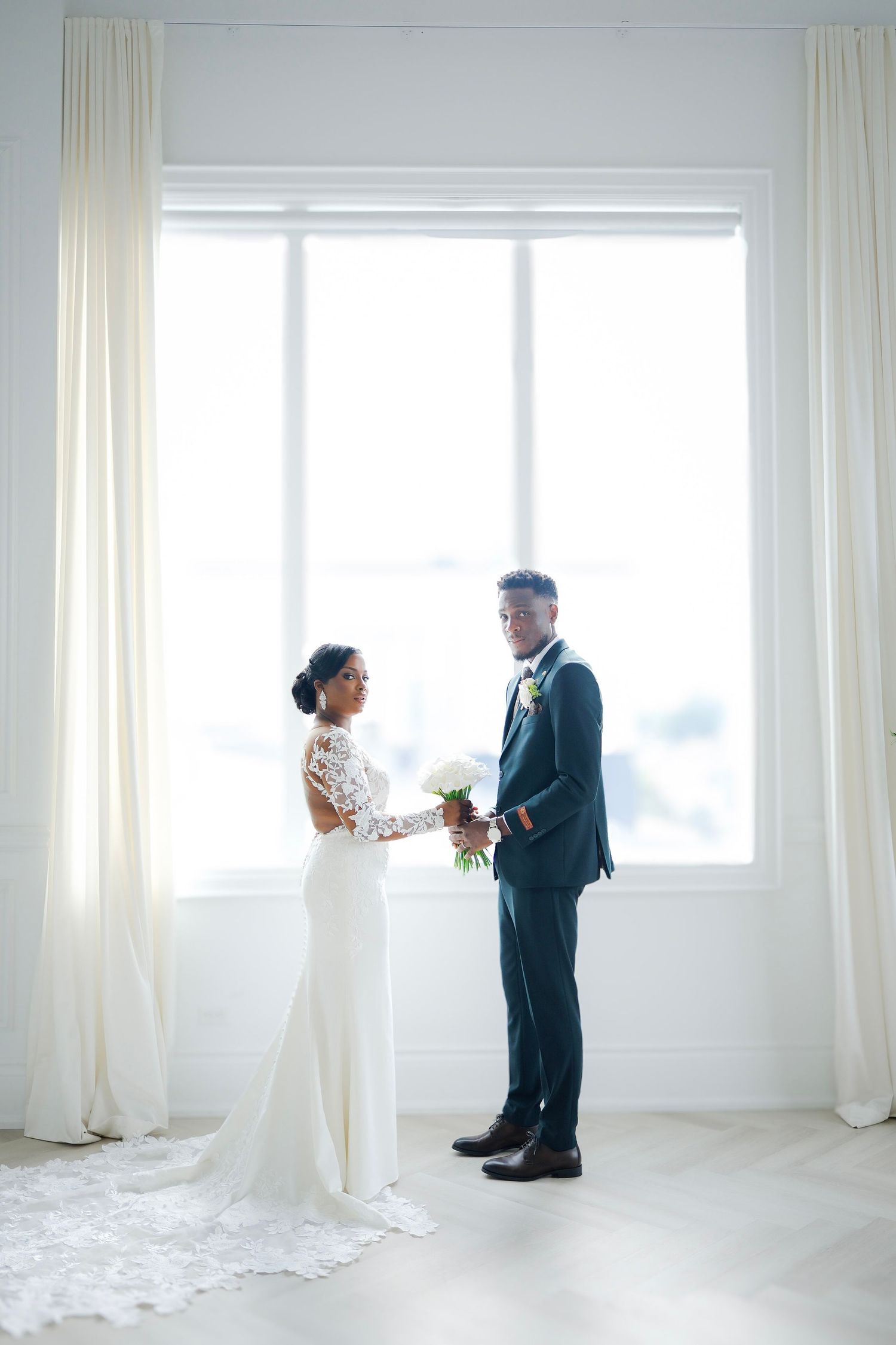 A couple in wedding attire stands face to face in front of bright window with flowing white curtains.