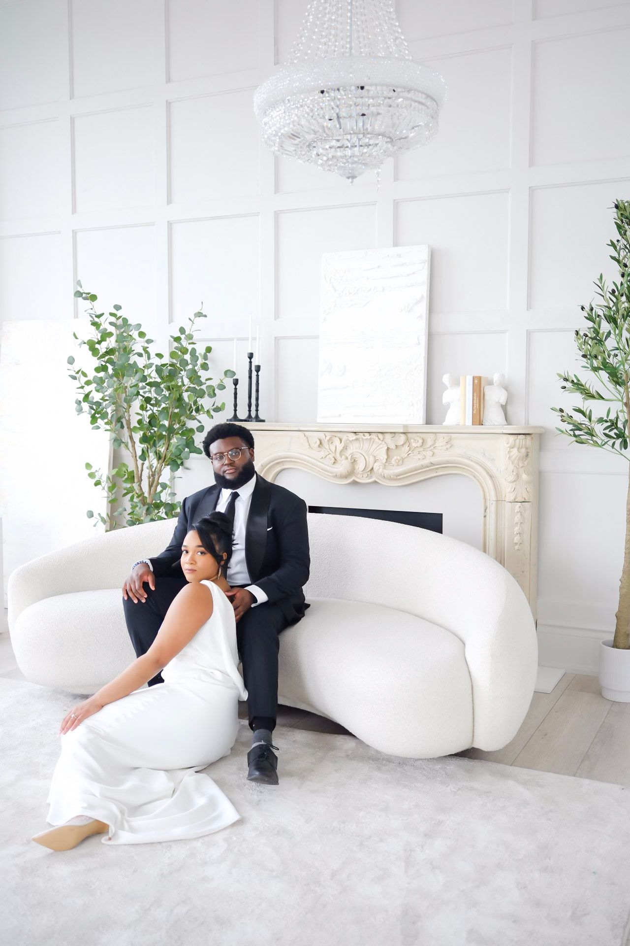 Elegant couple relaxes on curved white sofa in luxury room with ornate fireplace and crystal chandelier.