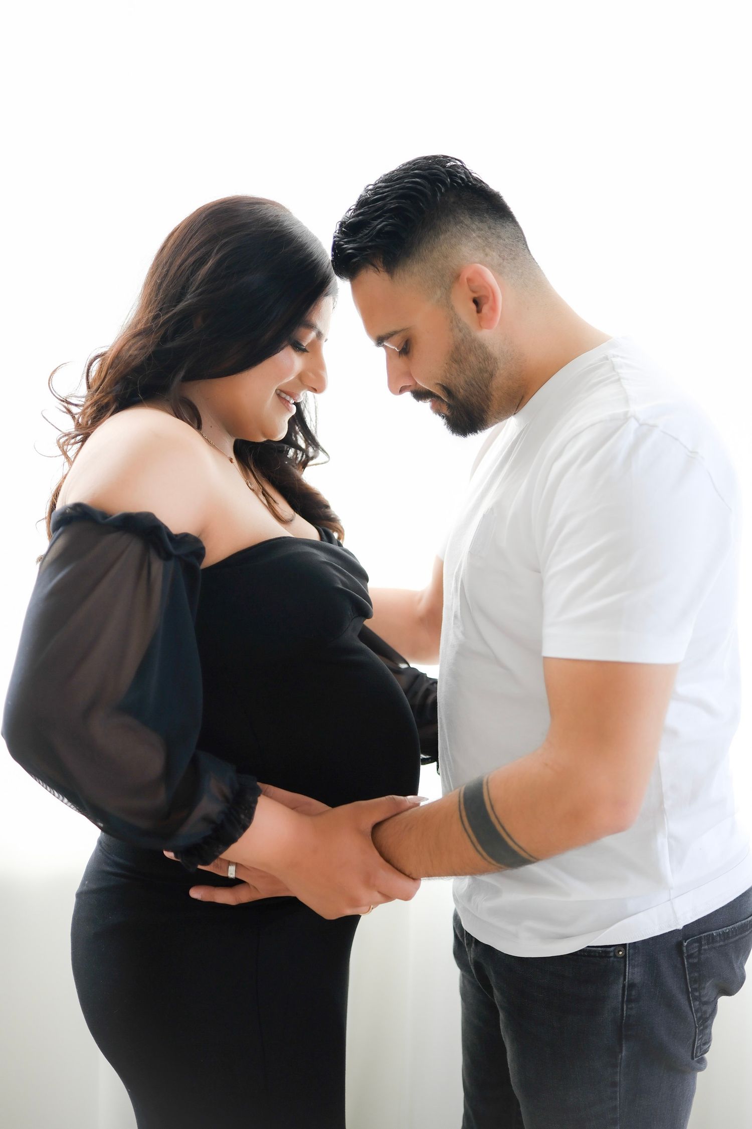 A couple in black and white outfits share an intimate moment during a maternity photoshoot against a light background.