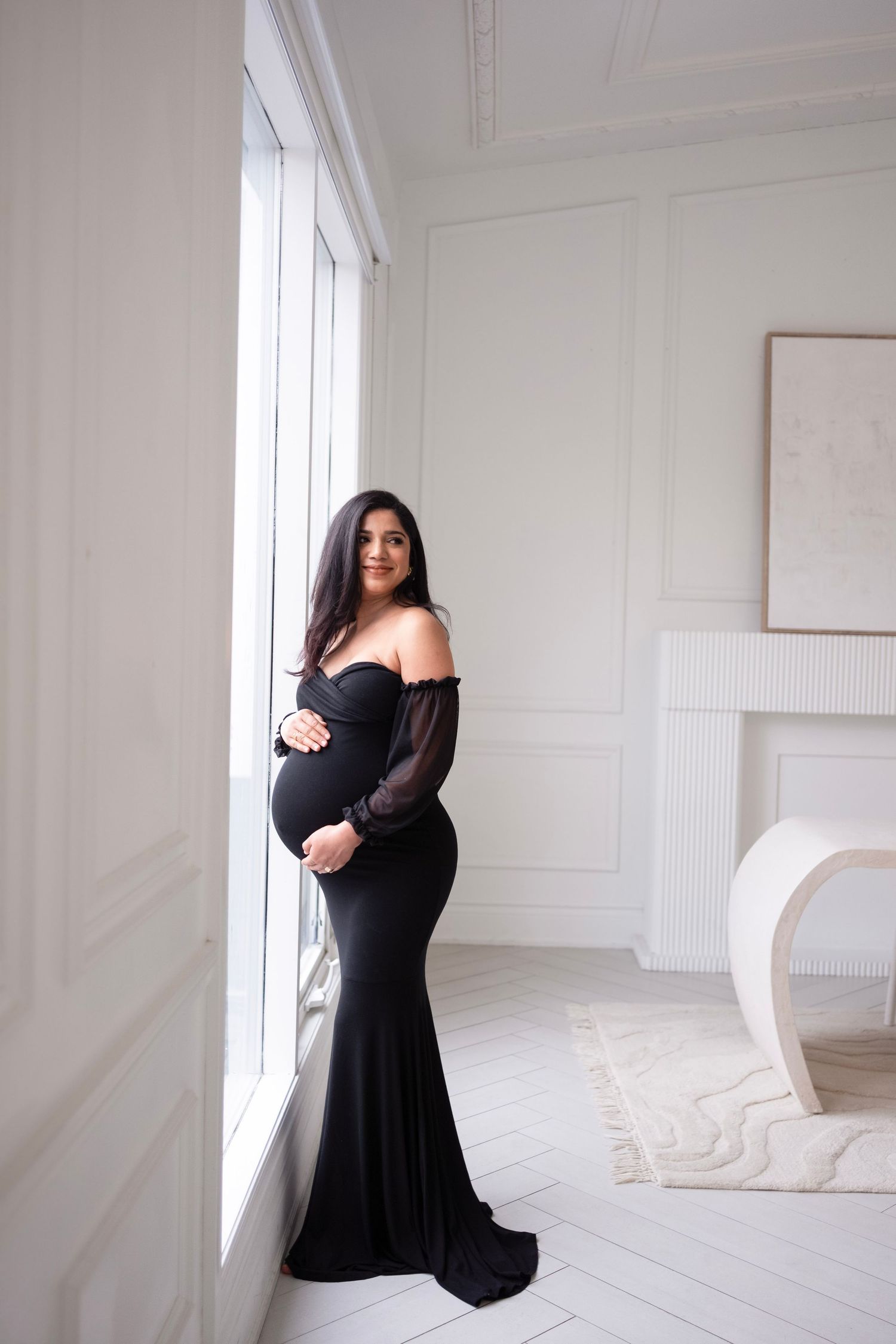Pregnant woman in elegant black off-shoulder gown standing by window in white room, cradling baby bump.