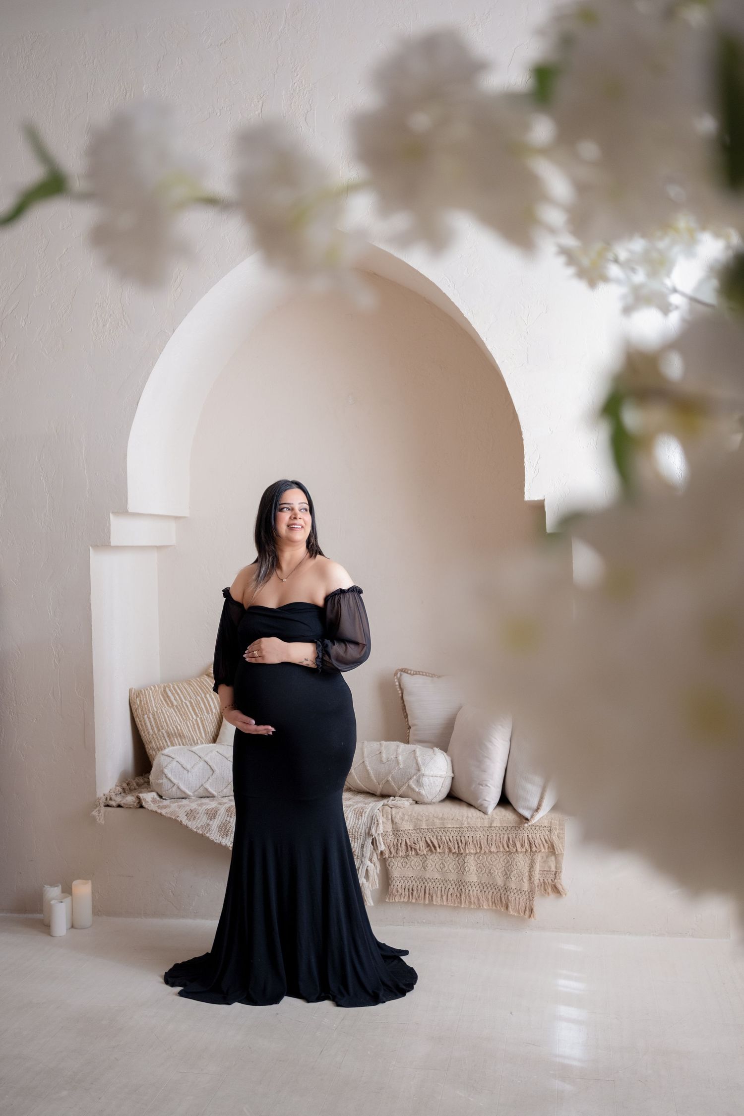 A maternity photo session in a bright white studio with soft floral elements framing a black dress against neutral tones.