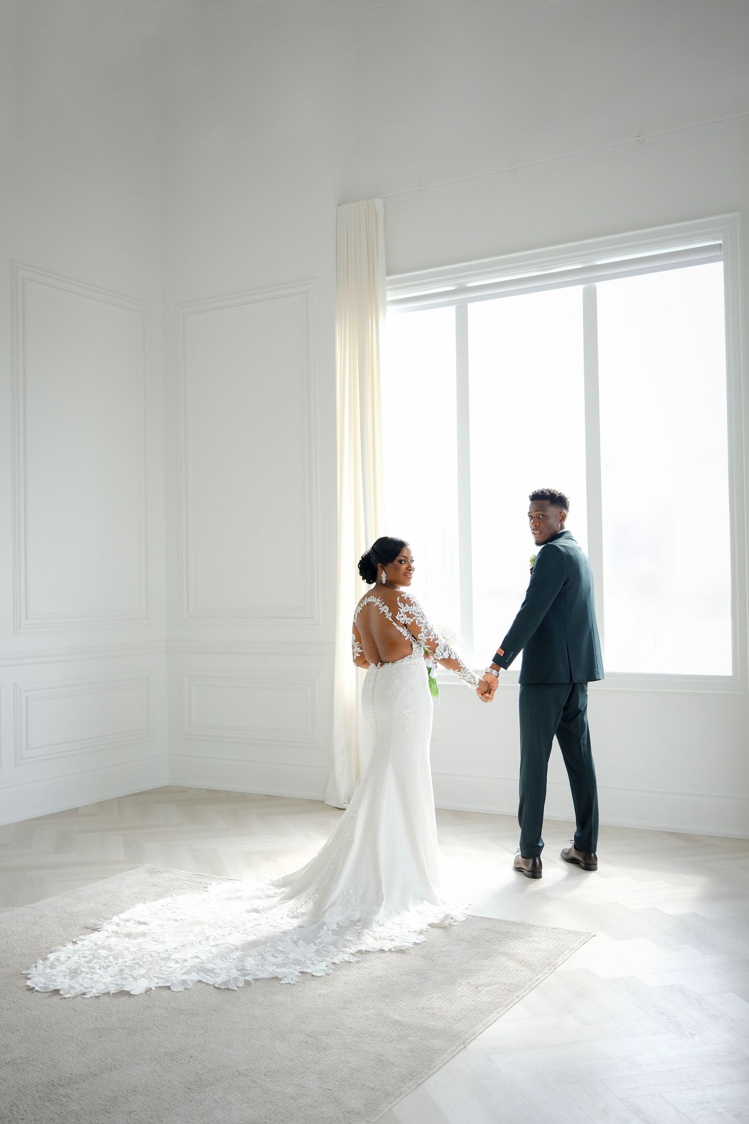 A couple in wedding attire stands together in an elegant minimalist white room with natural light streaming in through a window.