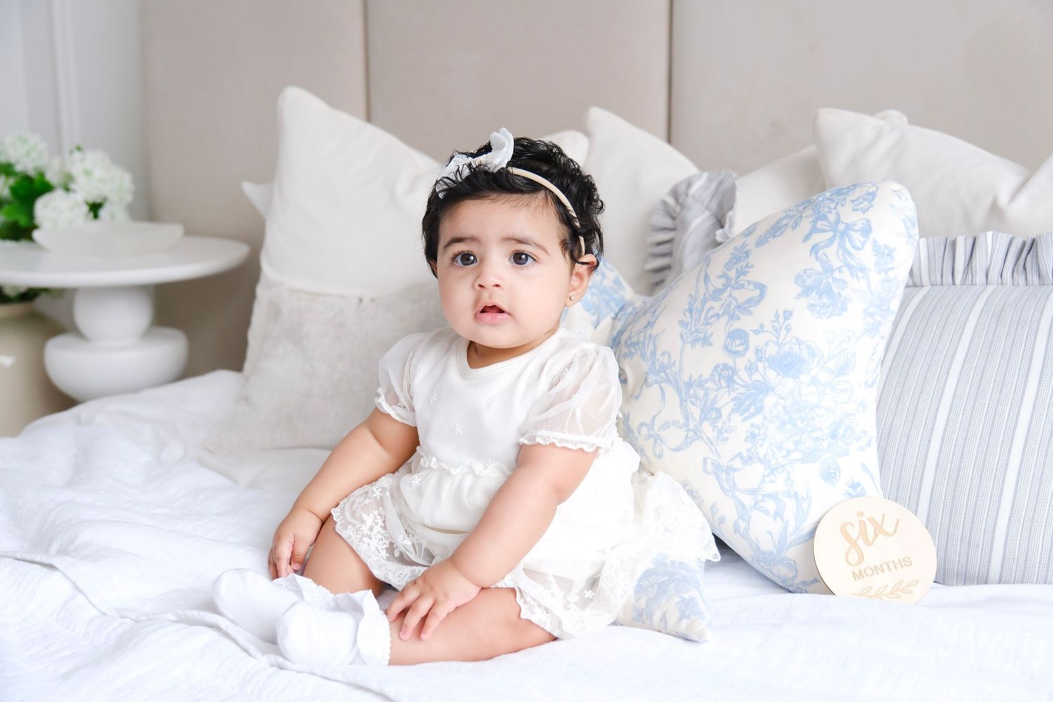 A baby wearing a white dress and headband sits on a cozy bed with light blue and white pillows.