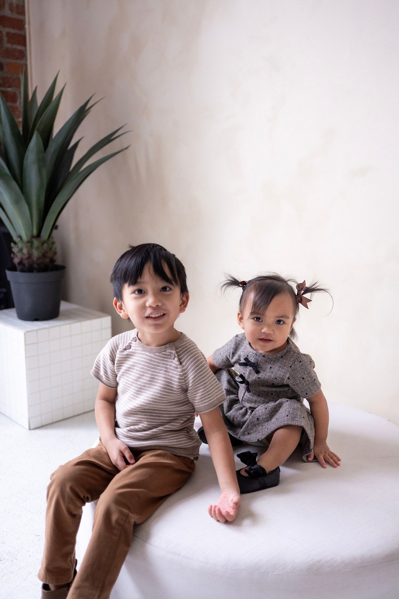 Two toddlers in matching striped shirts and brown pants sit together on a white surface near a potted plant.