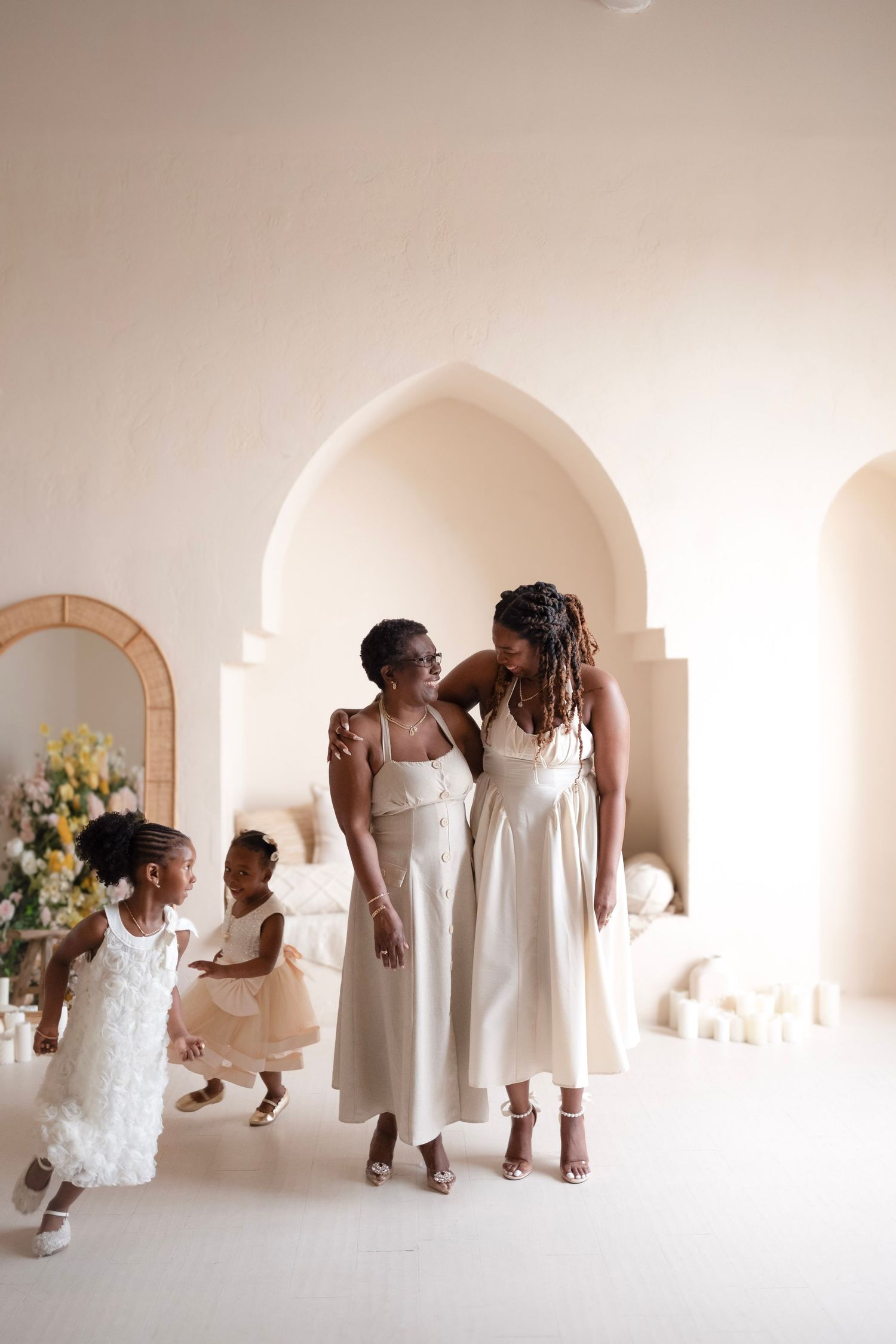 Two Black women in white midi dresses smile together as two young girls in white dresses play nearby in a bright, elegant room.