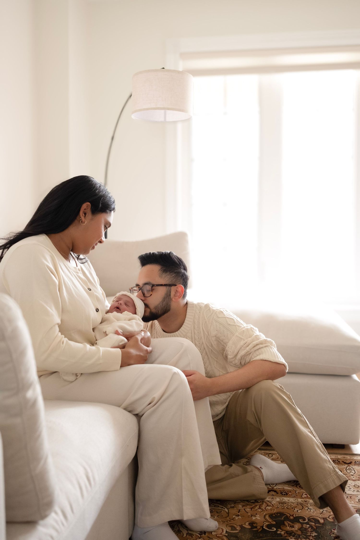 Young parents in neutral tones sitting on white sofa, lovingly gazing at their newborn baby in a bright living room.