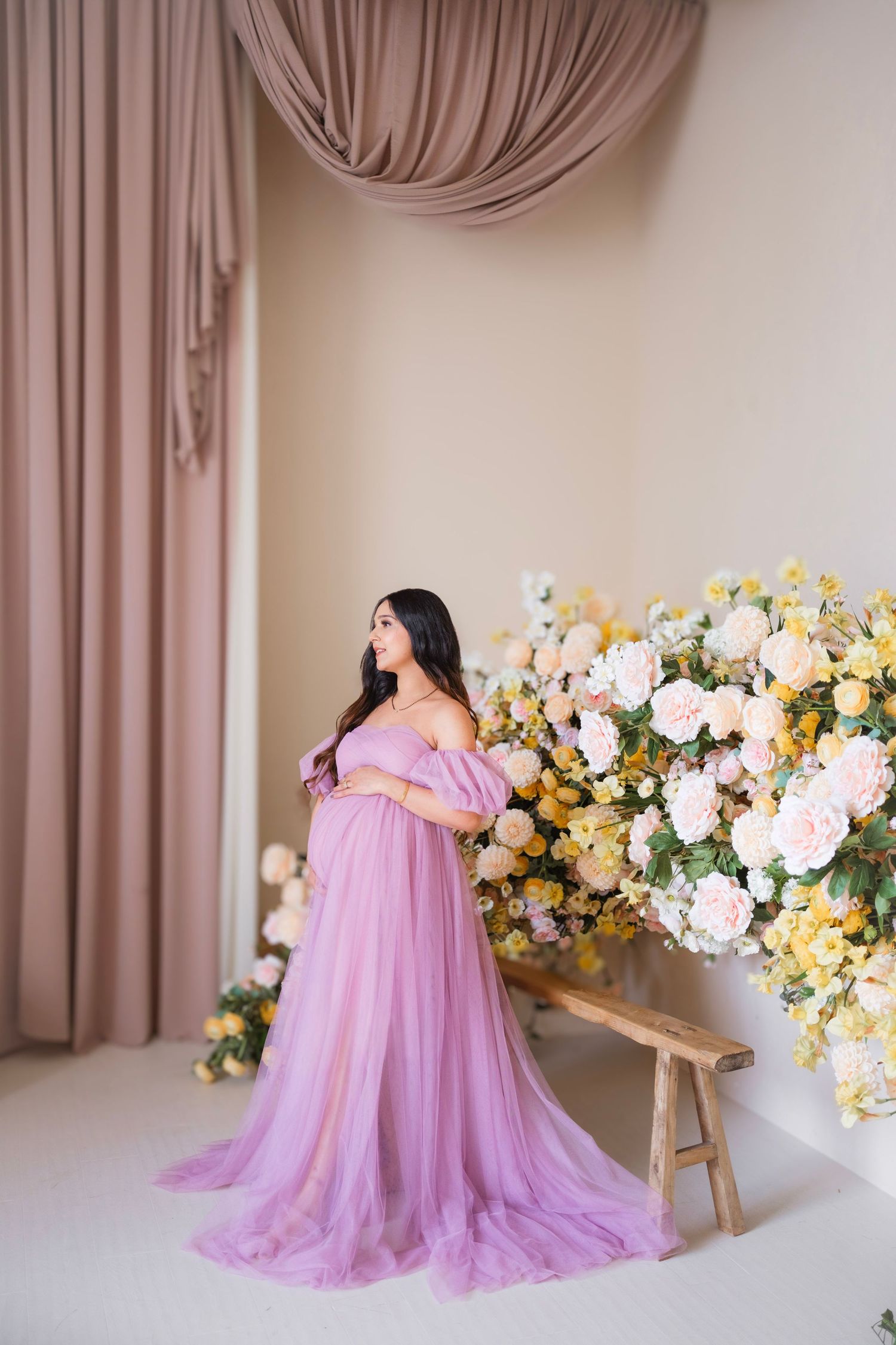Pregnant woman in lavender tulle gown poses gracefully beside floral arrangement in elegant studio setting.