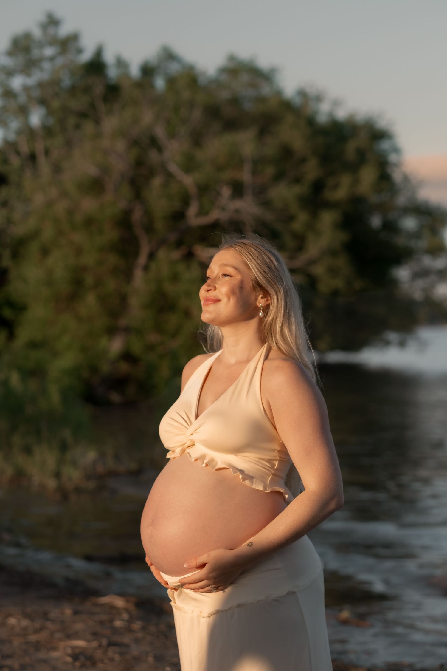 Pregnant woman in cream outfit cradling her baby bump outdoors by a lake at golden hour sunset.