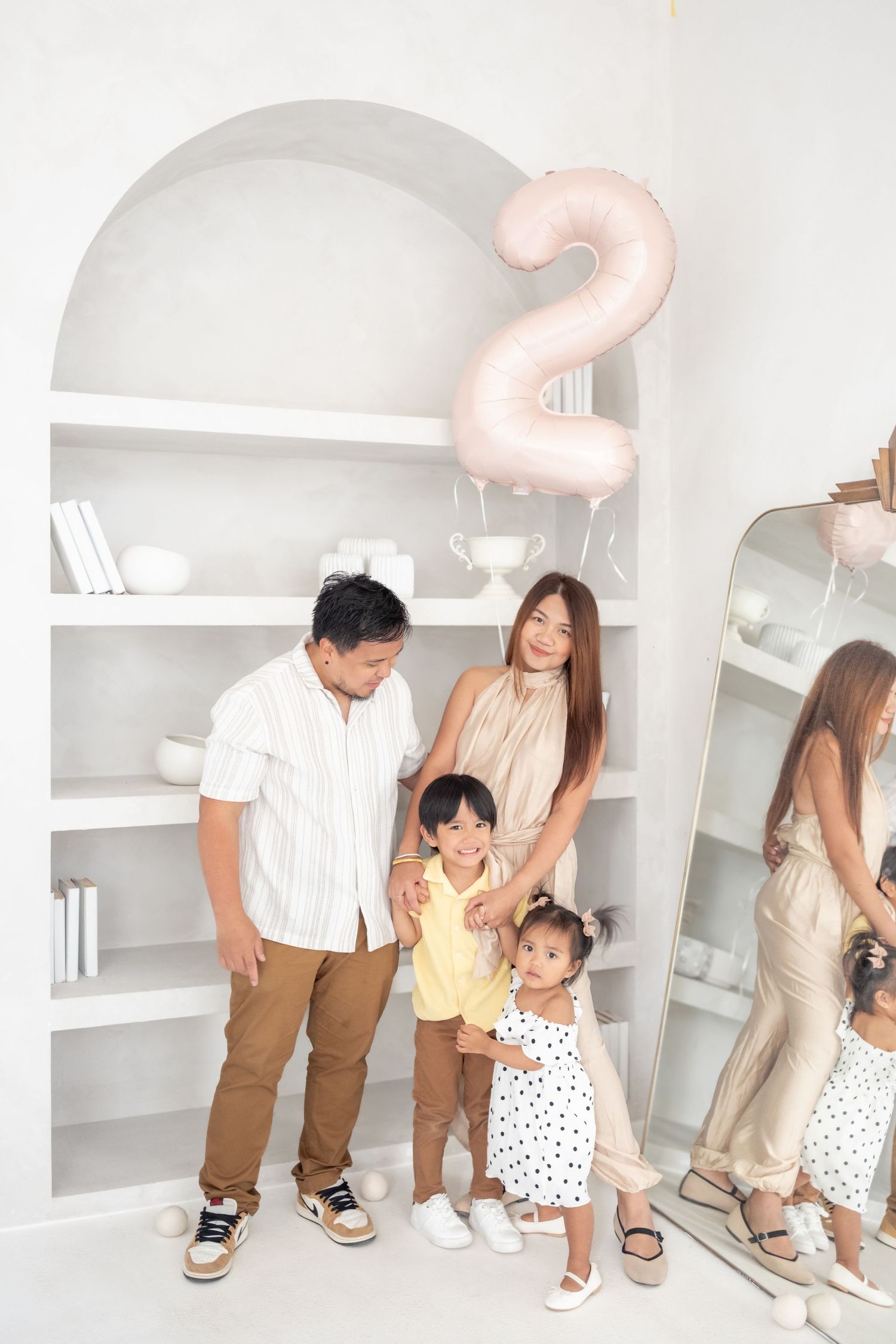 Asian family of four celebrating a 2nd birthday with a pink number balloon in a bright white room with shelving.