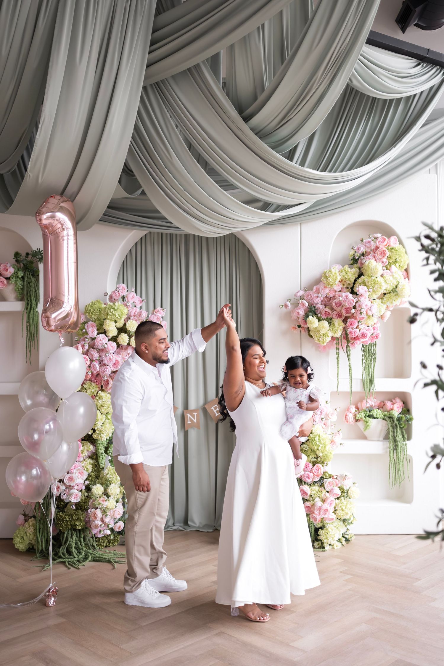 Family celebrates baby's first birthday in elegant venue with sage green drapes, pink floral arrangements and balloons.