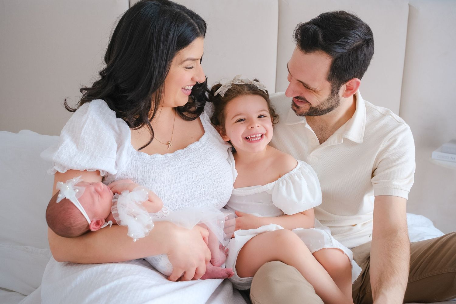 Happy family sitting together on white bed at home with newborn baby and young child smiling.