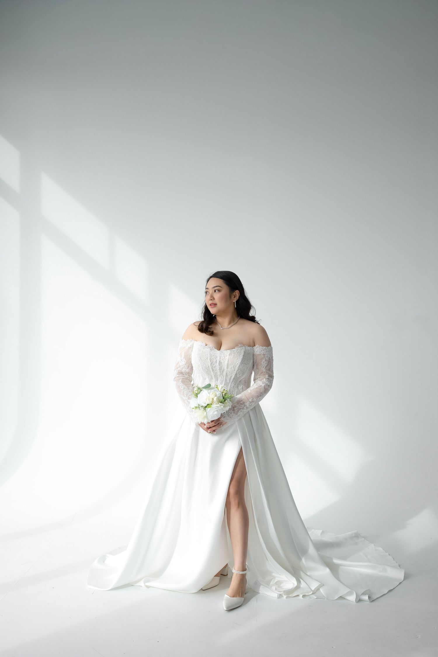 Elegant bride in white off-shoulder lace wedding gown with side slit holds bouquet in sunlit white studio.