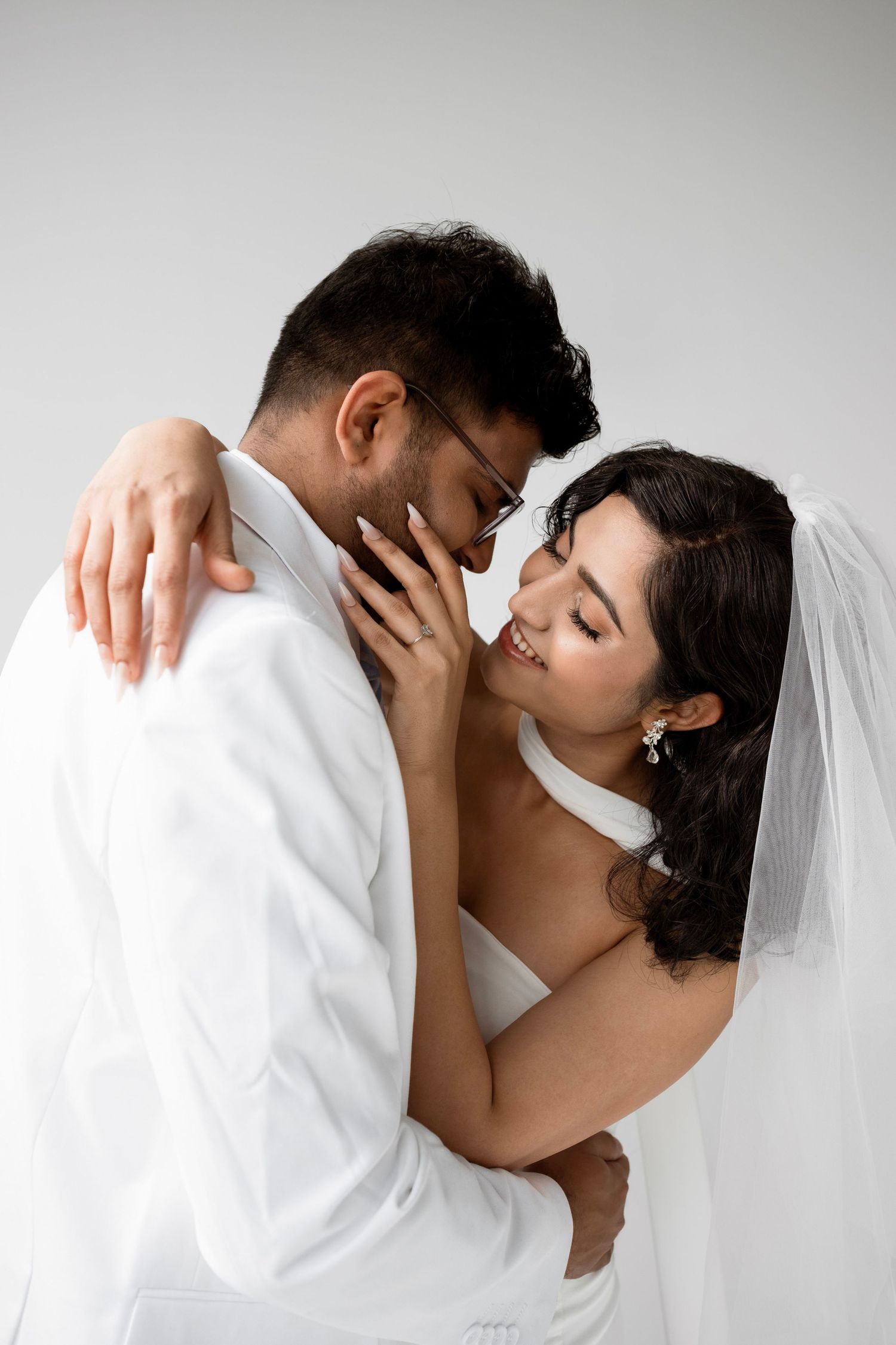 A bride and groom in white wedding attire share an intimate moment, smiling close together against a grey background.