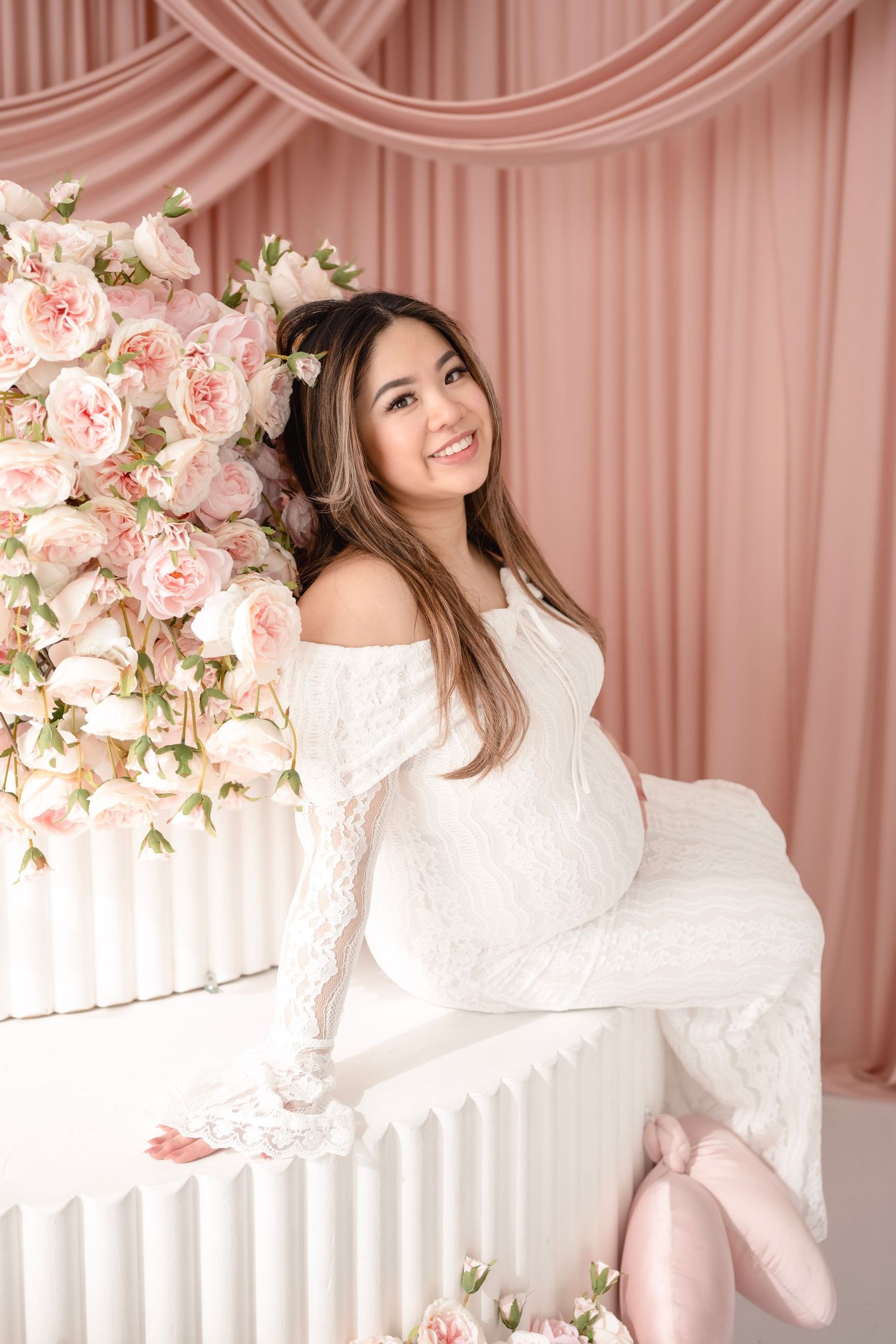 Smiling pregnant woman in white lace off-shoulder gown seated among pink roses against blush pink draped backdrop.