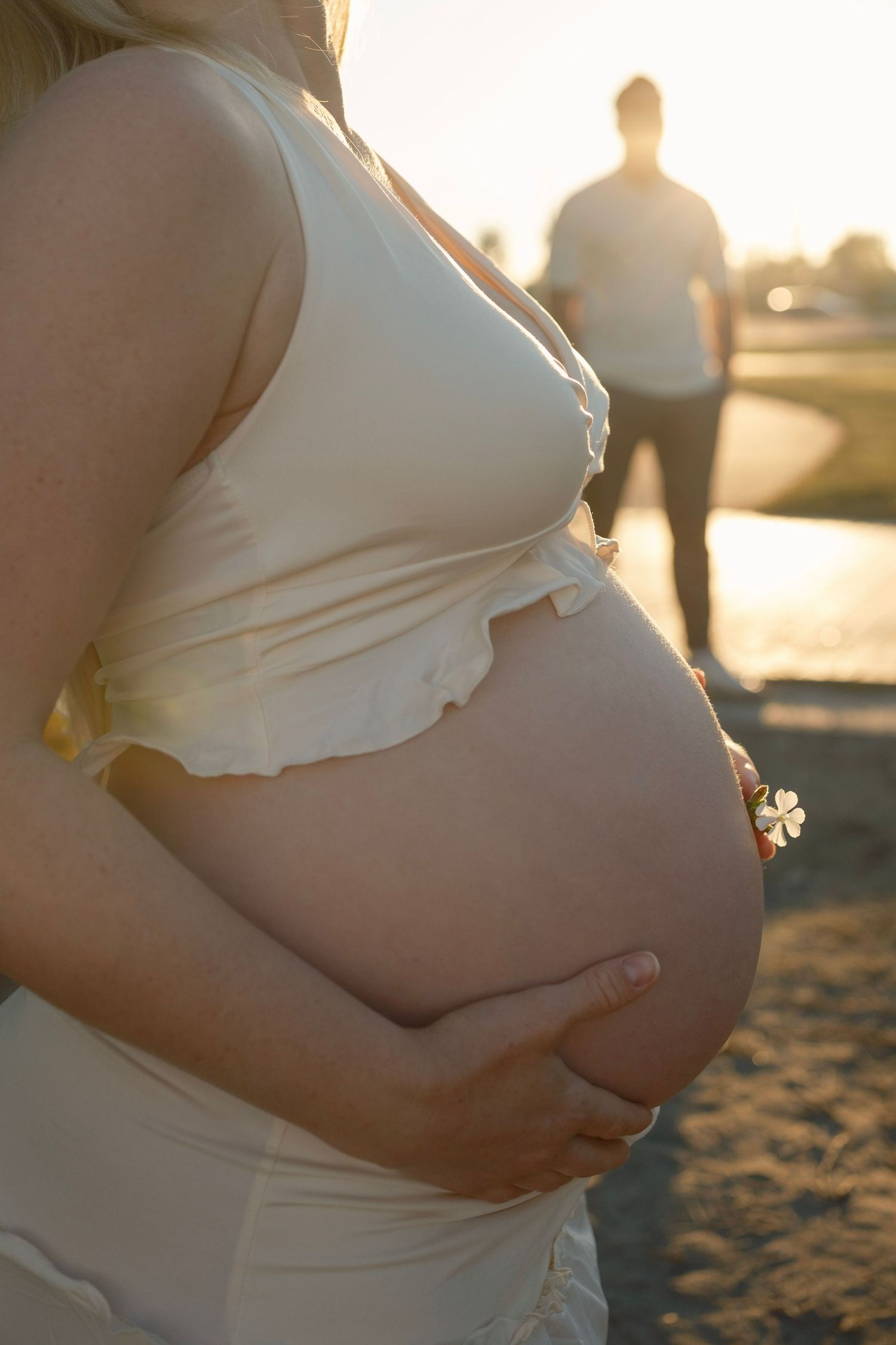 Pregnant woman in white dress cradling baby bump at golden sunset with man standing in background.