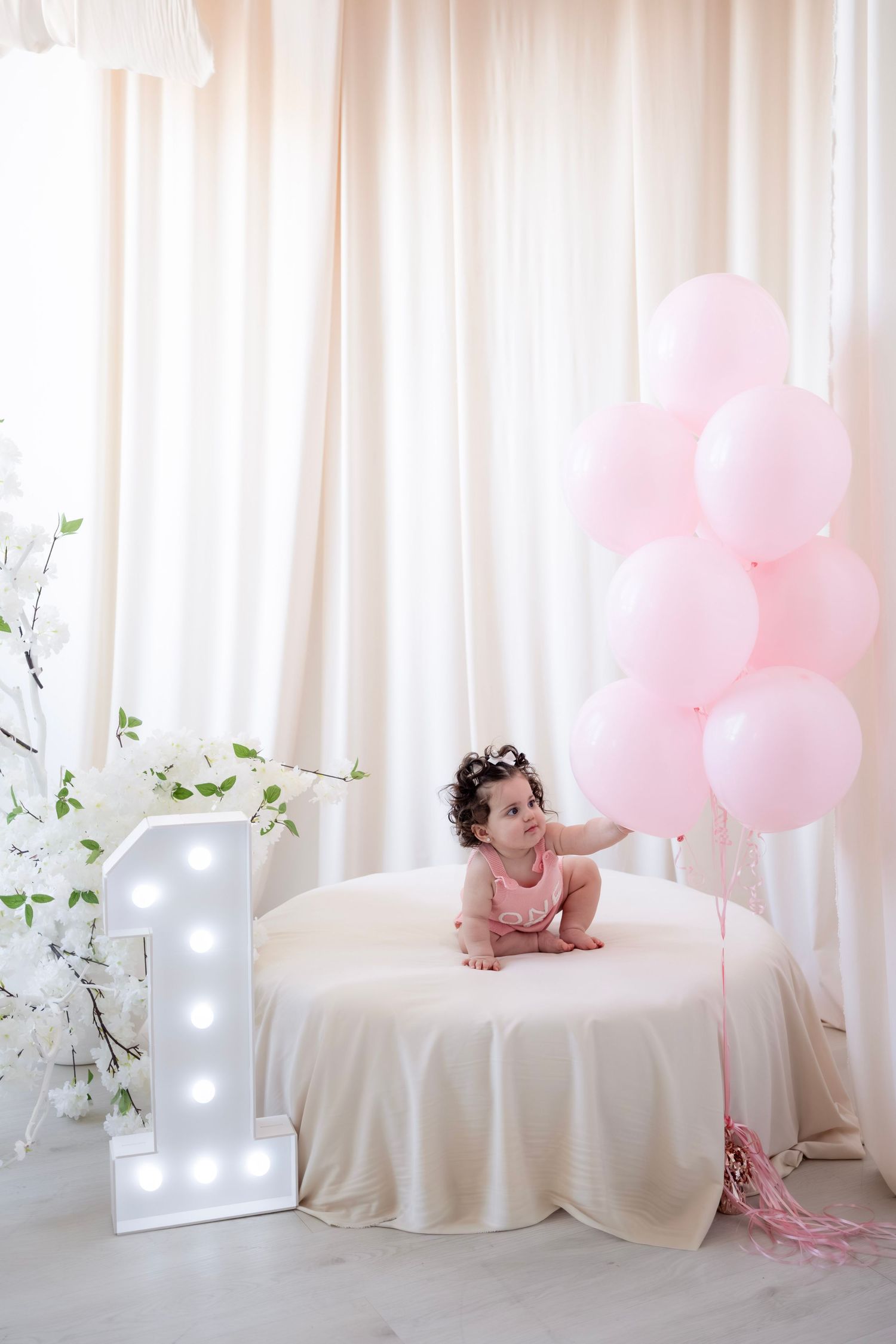 Adorable baby girl in pink romper celebrating first birthday with lit number one sign and pink balloons.