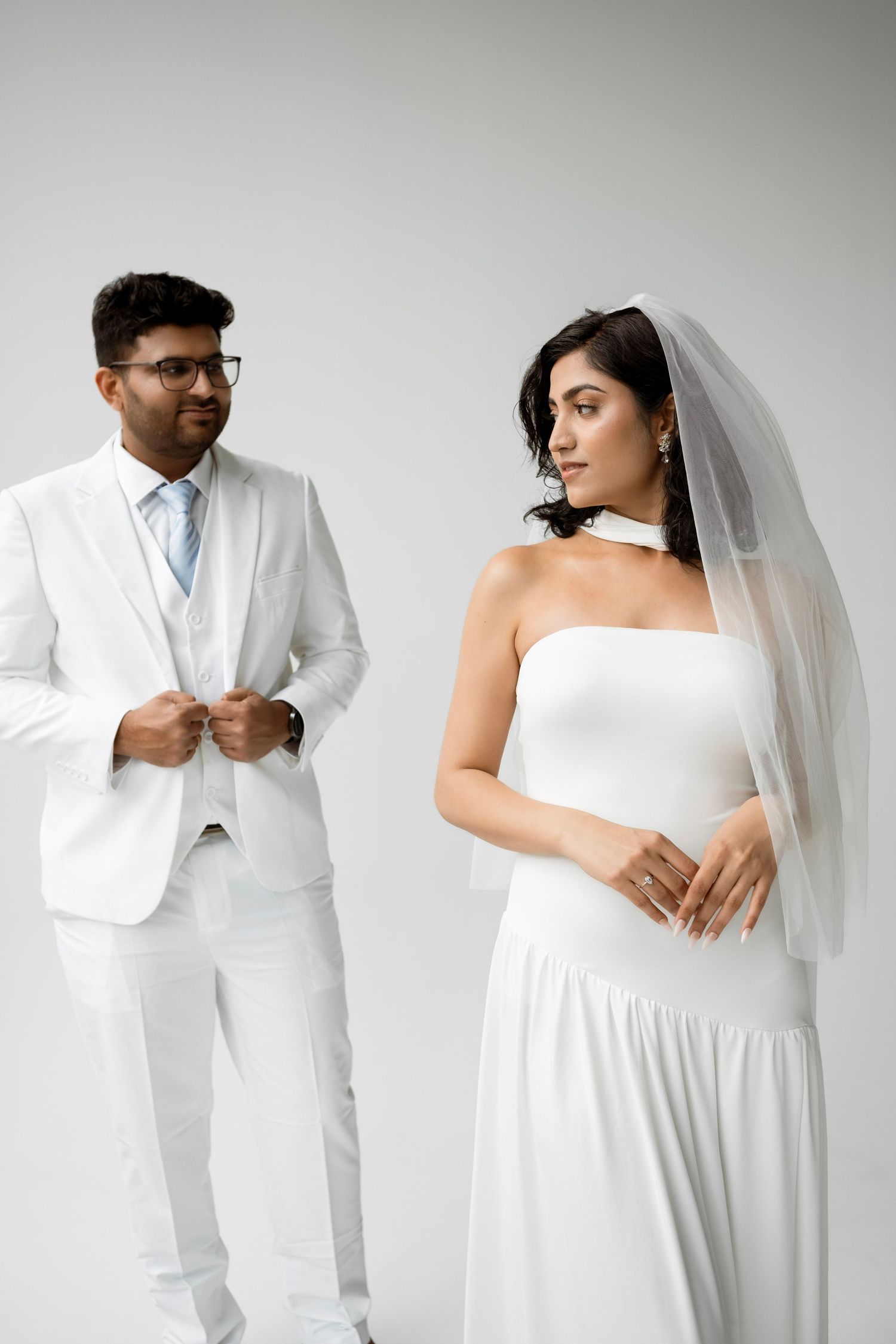Indian bride in white strapless gown with veil stands beside groom in white suit on light background.